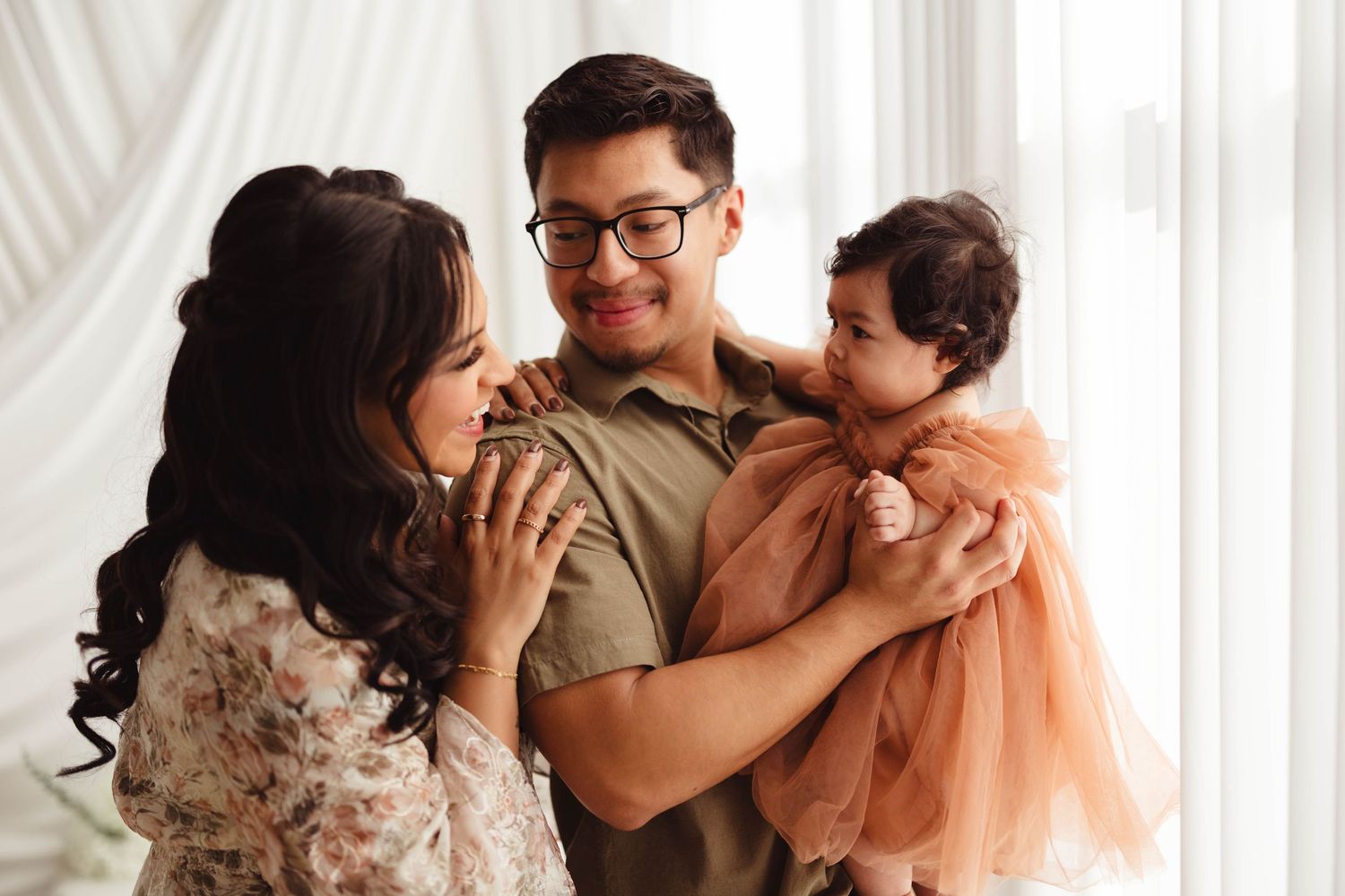 Happy family with parents and baby girl in elegant outfits posing together near bright window. Lafayette indiana tippecanoe county west lafa