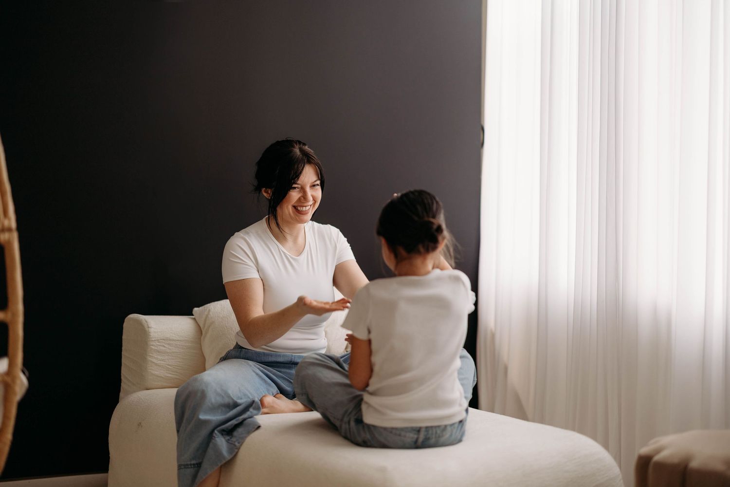 Dark haired mother laughing and playing with young daughter on white sofa against dark wall. Motherhood  in west Lafayette indiana