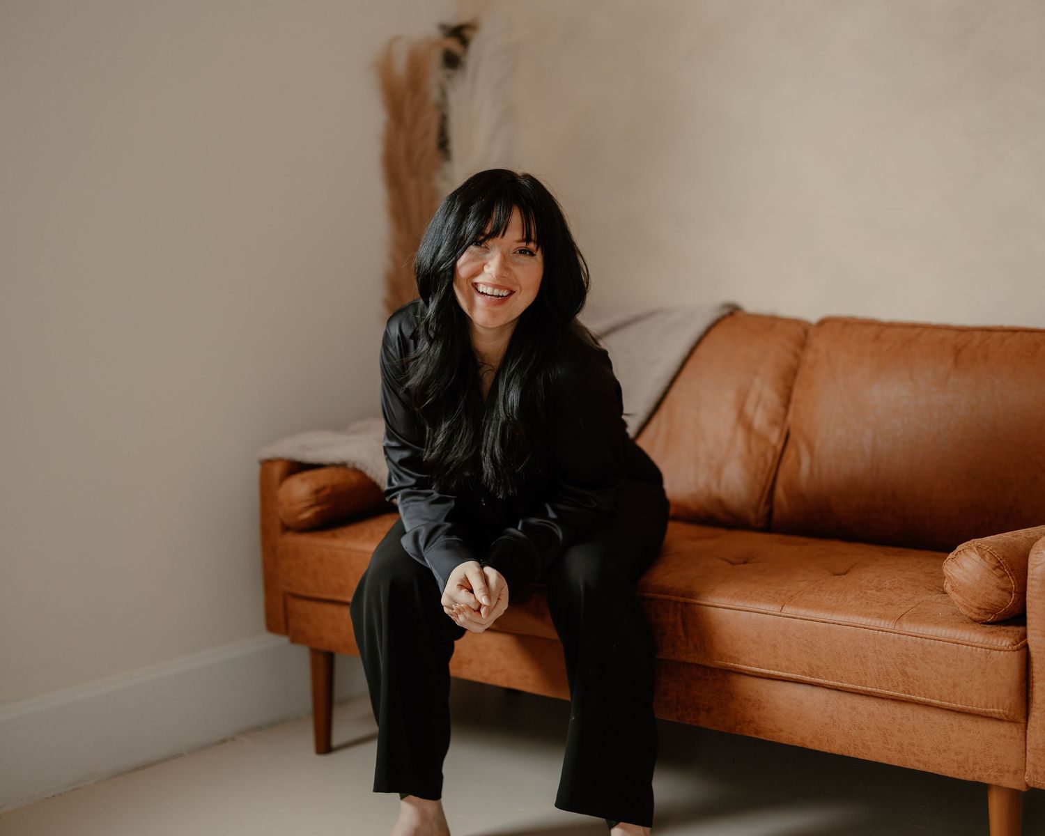 Smiling young woman in black outfit sitting on edge of brown leather couch. Branding photos woman owned business in west Lafayette Indiana