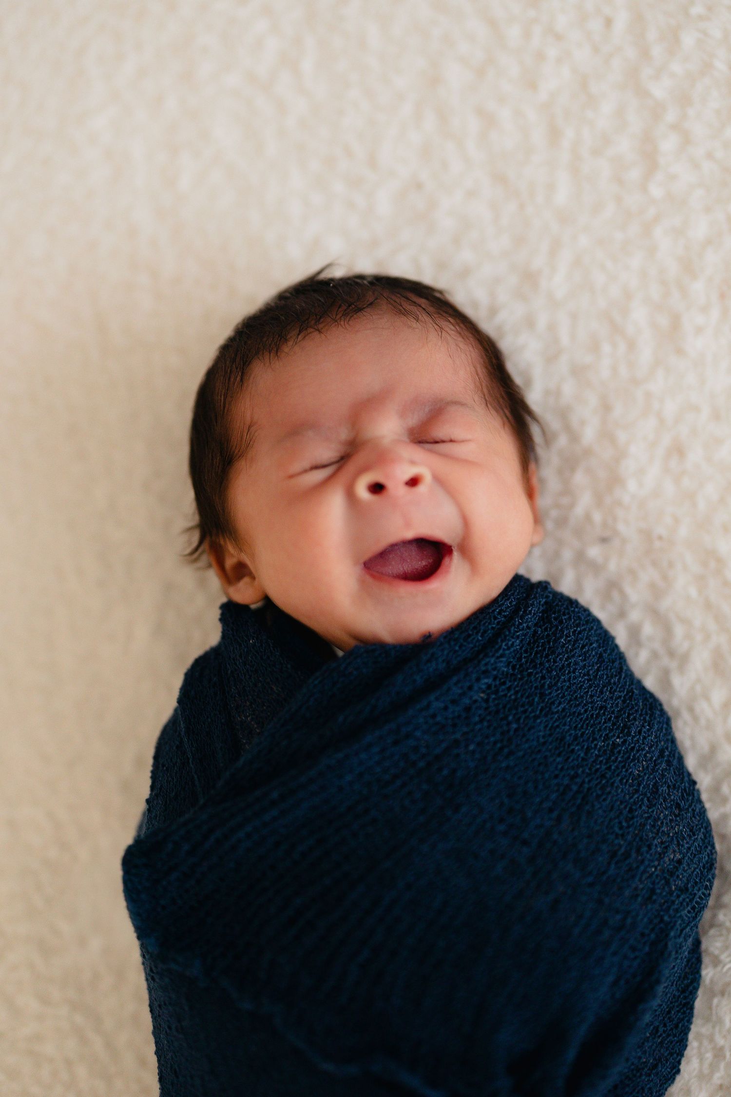 Newborn baby swaddled in dark blue knit wrap yawning widely while lying on white fluffy surface. Lafayette indiana tippecanoe county
