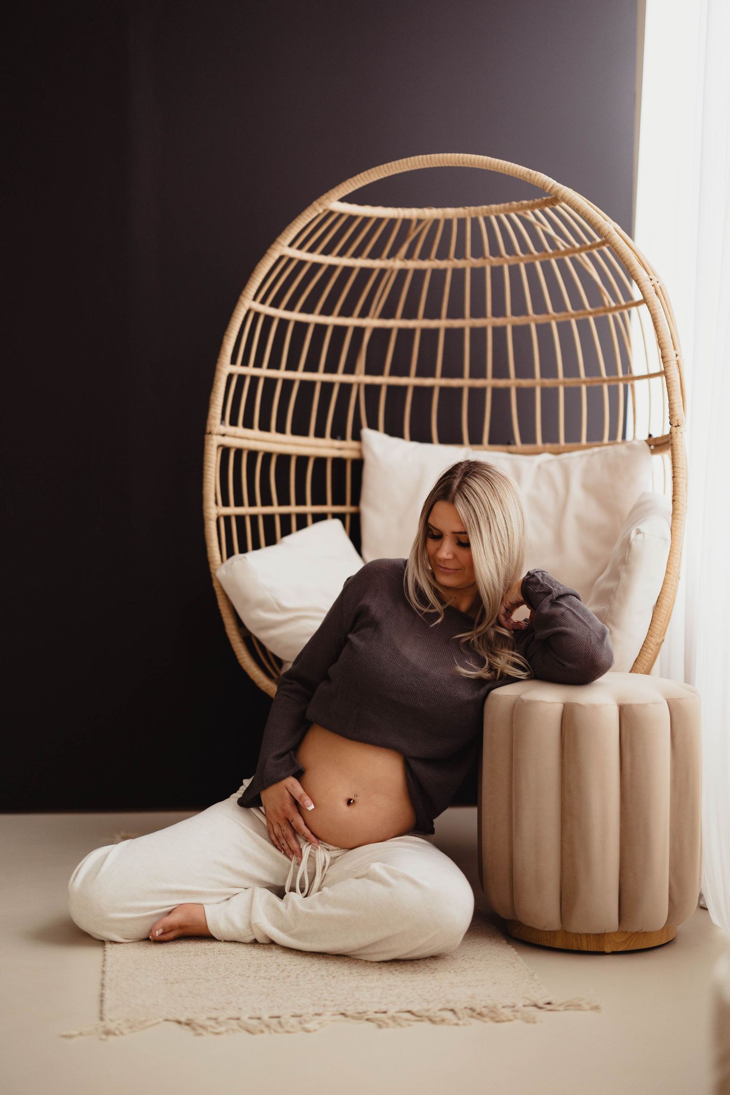 Pregnant blonde woman sitting by rattan egg chair touching bare belly indoors. Lafayette indiana tippecanoe county west lafayette