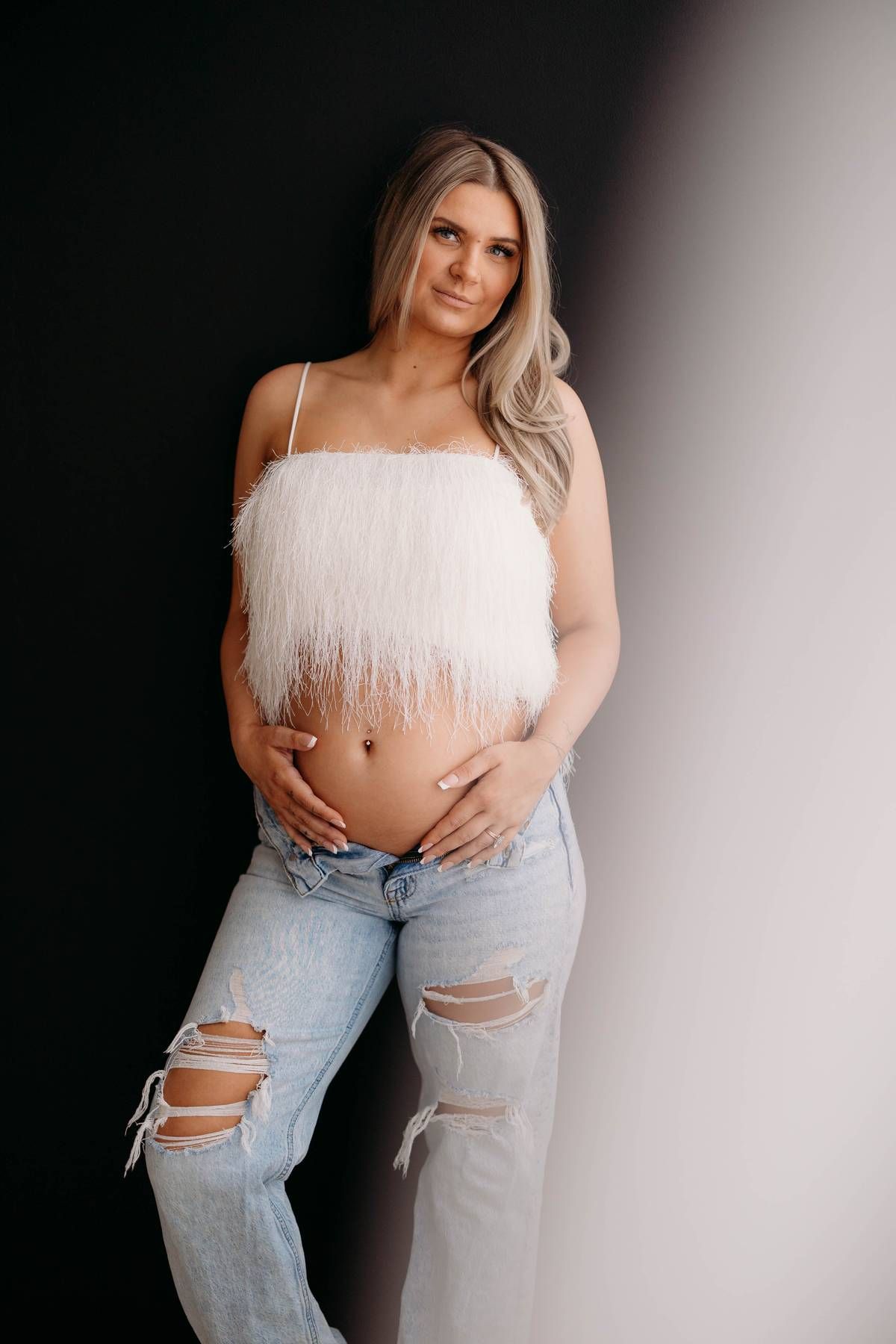 Pregnant woman in white fluffy crop top and ripped jeans cradling baby bump against black and white background. maternity Lafayette indiana