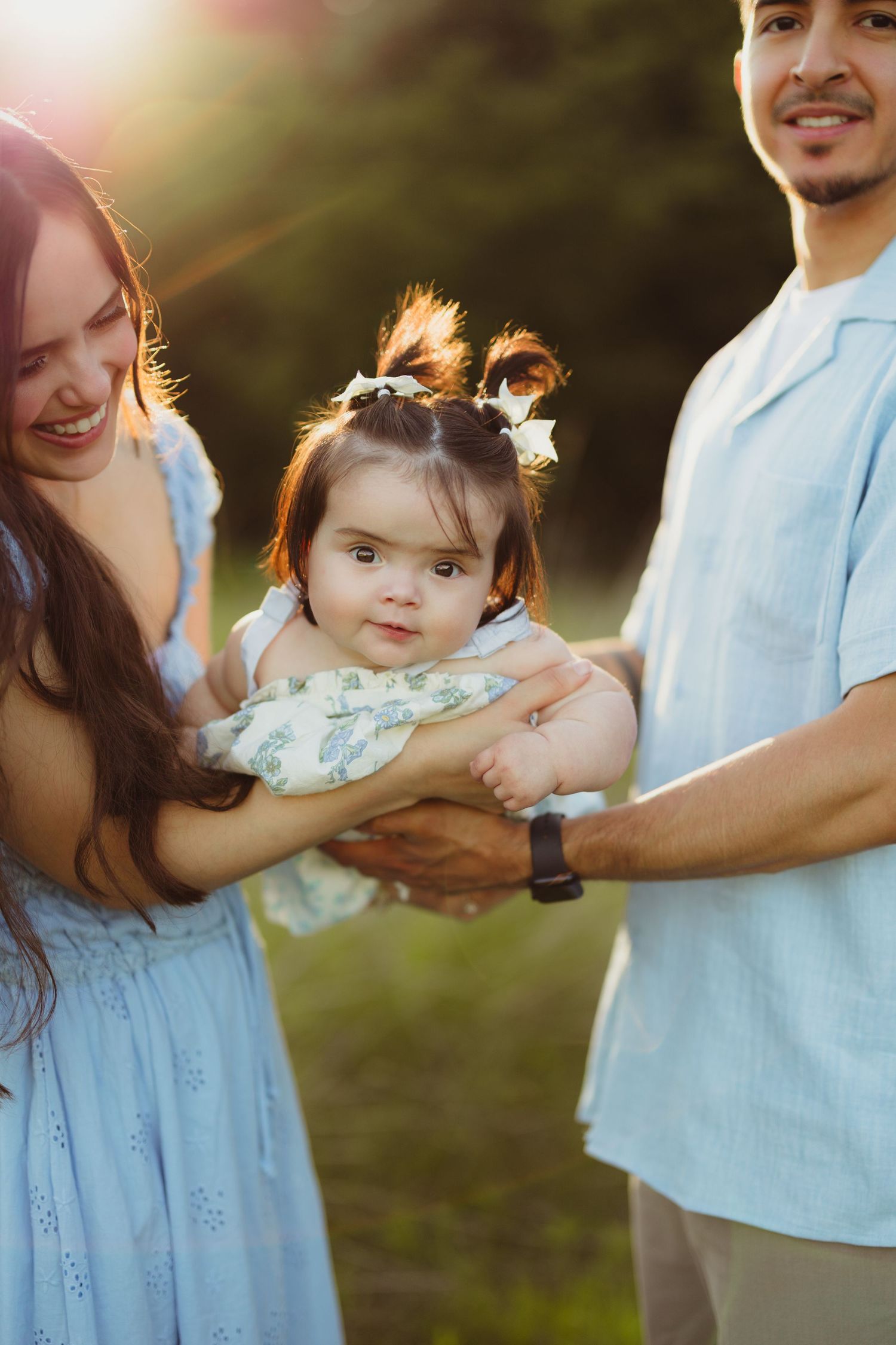 A young family with a baby girl enjoys a golden hour outdoor photoshoot, parents in matching light blue outfits.
