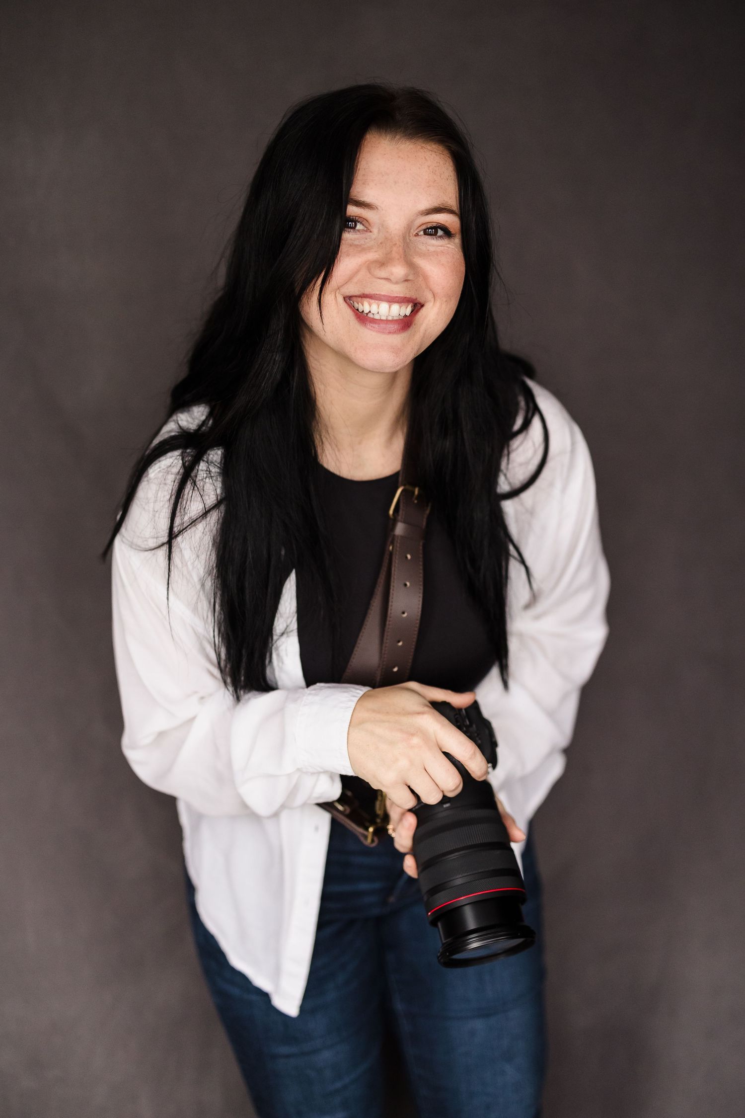 Smiling female photographer with long dark hair holding a camera in a studio setting. Lafayette indiana tippecanoe county west lafayette