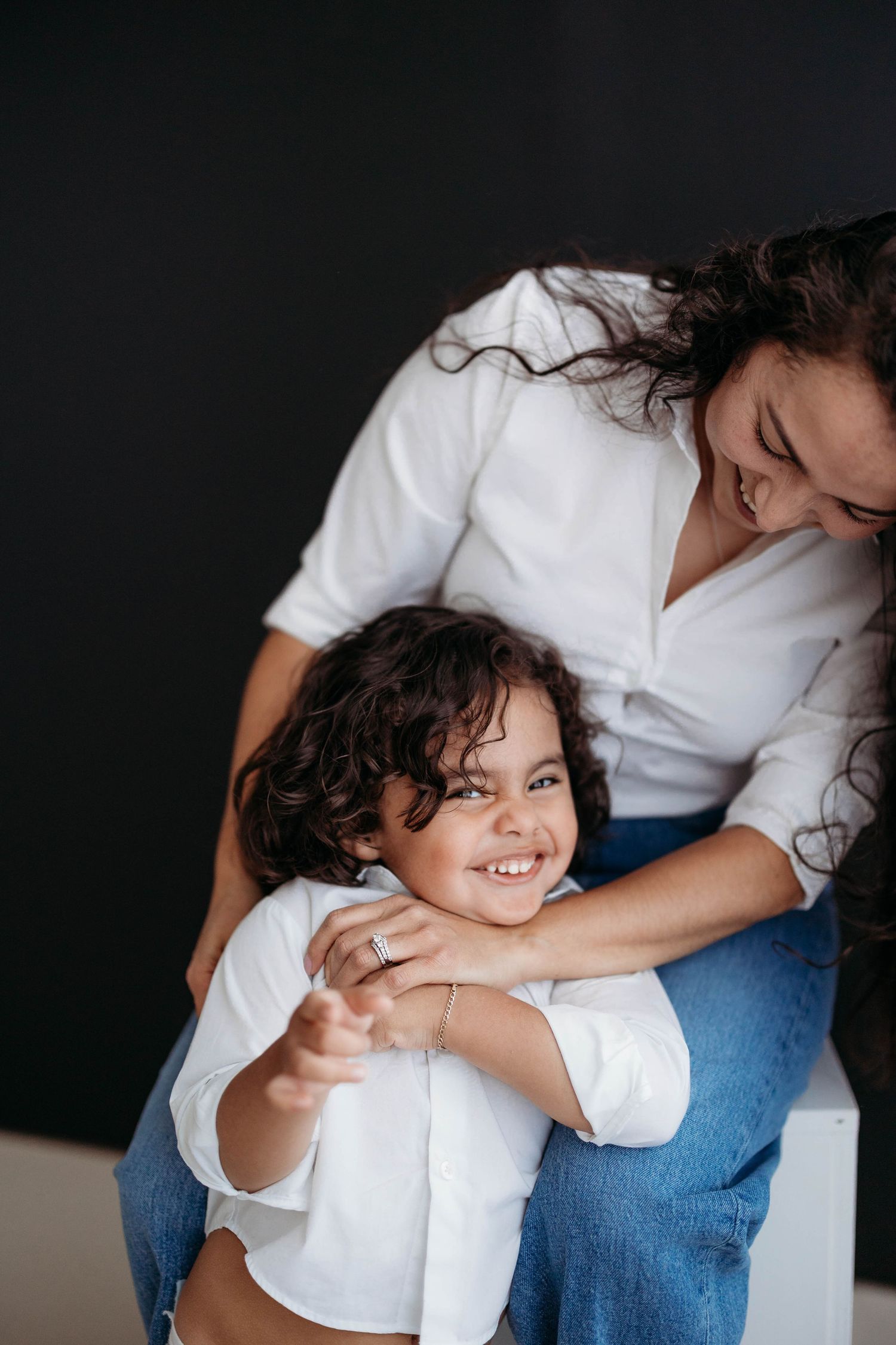 Smiling mother embracing her happy curly-haired child sitting on white cube in studio. Lafayette indiana tippecanoe county west lafayette