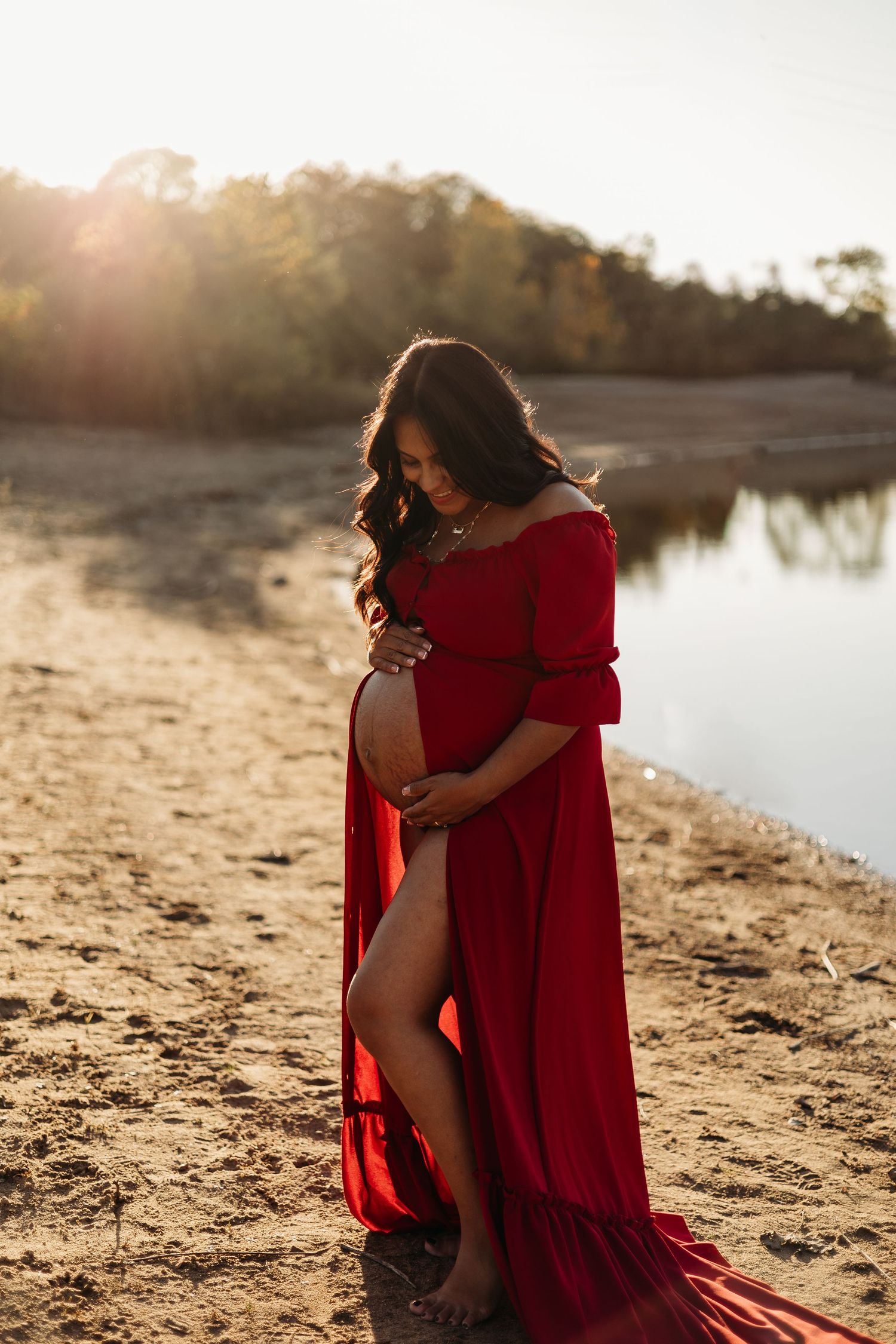 Pregnant woman in flowing red dress with slit cradling bump on sandy riverbank at golden hour. Lafayette indiana tippecanoe county