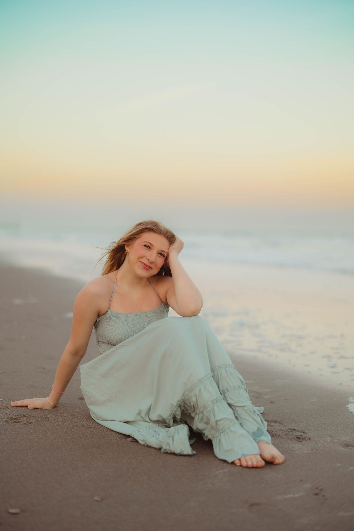 Blonde woman in mint green dress sitting on wet sand by ocean shore at sunset. High school senior photos in west Lafayette indiana