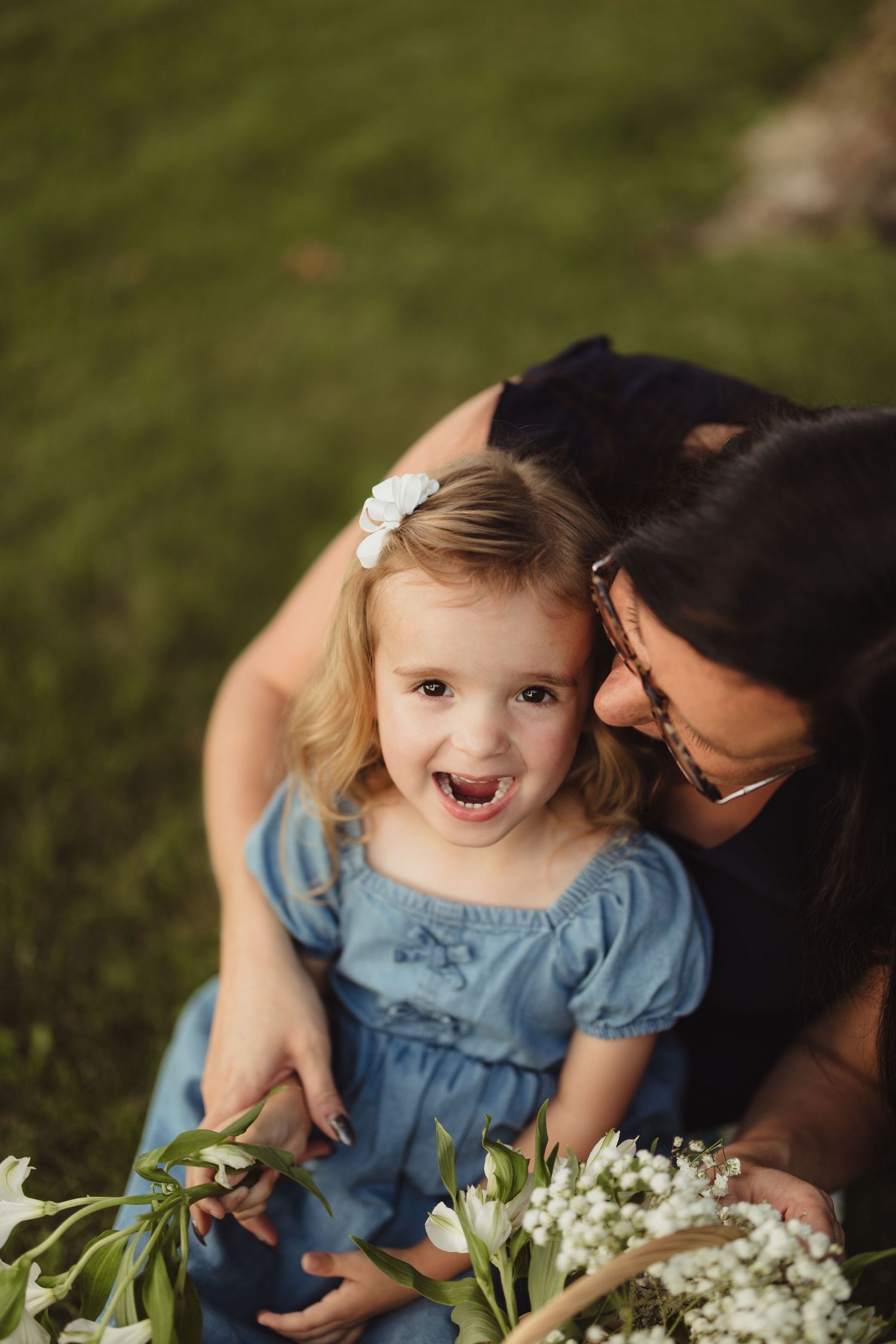 A mother kisses her smiling toddler girl in a blue dress holding white flowers on green grass.