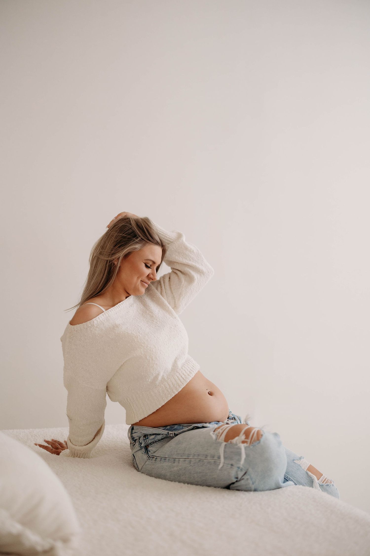 Blonde pregnant woman in white sweater and jeans sitting on bed admiring bump. Lafayette indiana tippecanoe county west lafayette