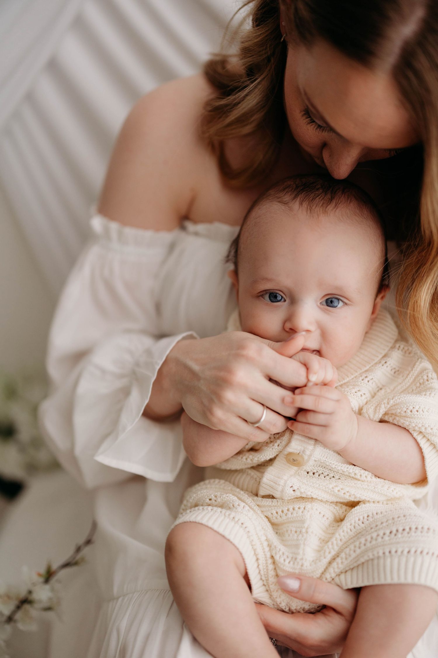 Mother in white off-shoulder dress cradles blue-eyed baby chewing fingers in bright studio. Lafayette indiana west Lafayette indiana