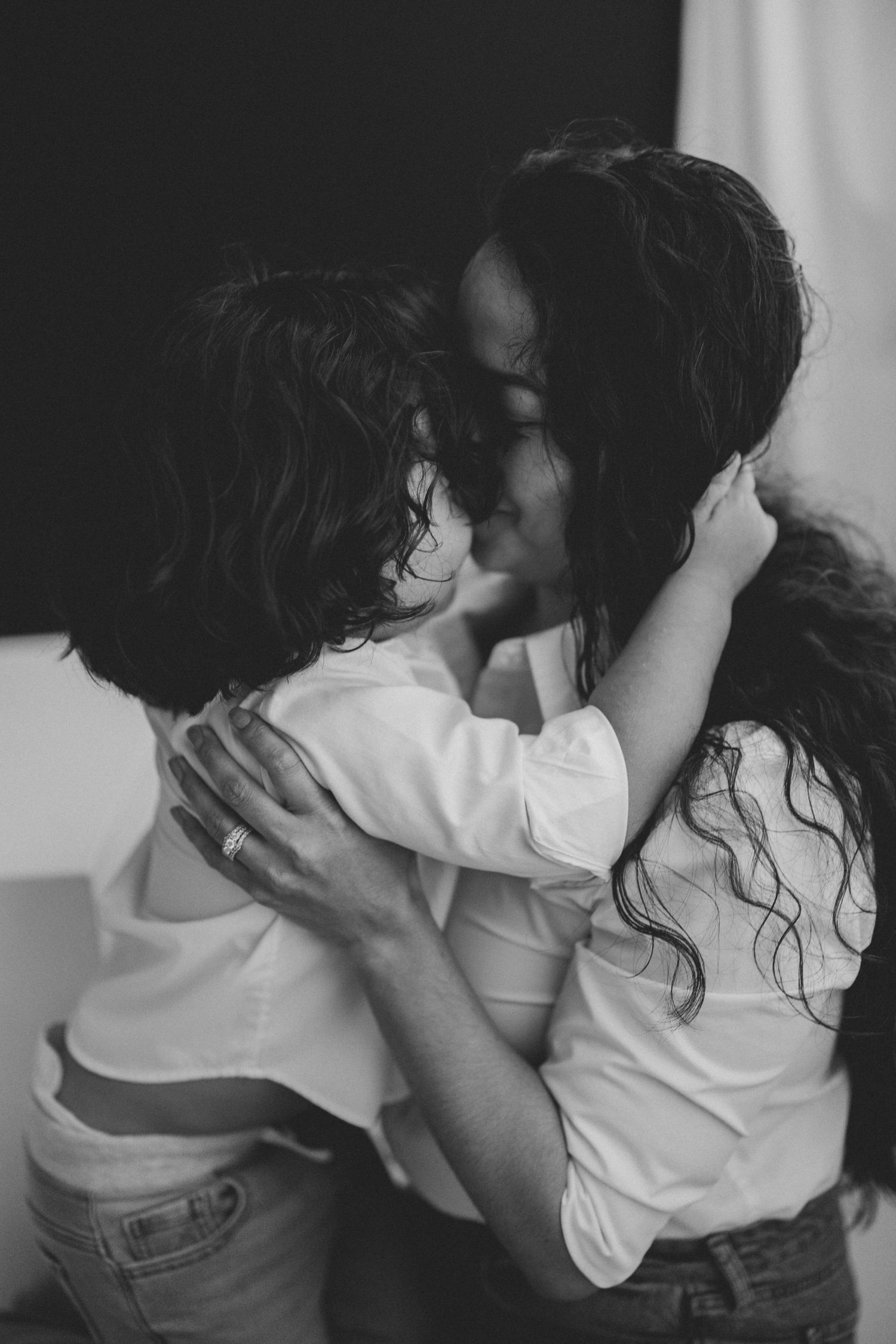 Black and white photo of two women with curly hair embracing and kissing, both wearing white shirts and jeans.