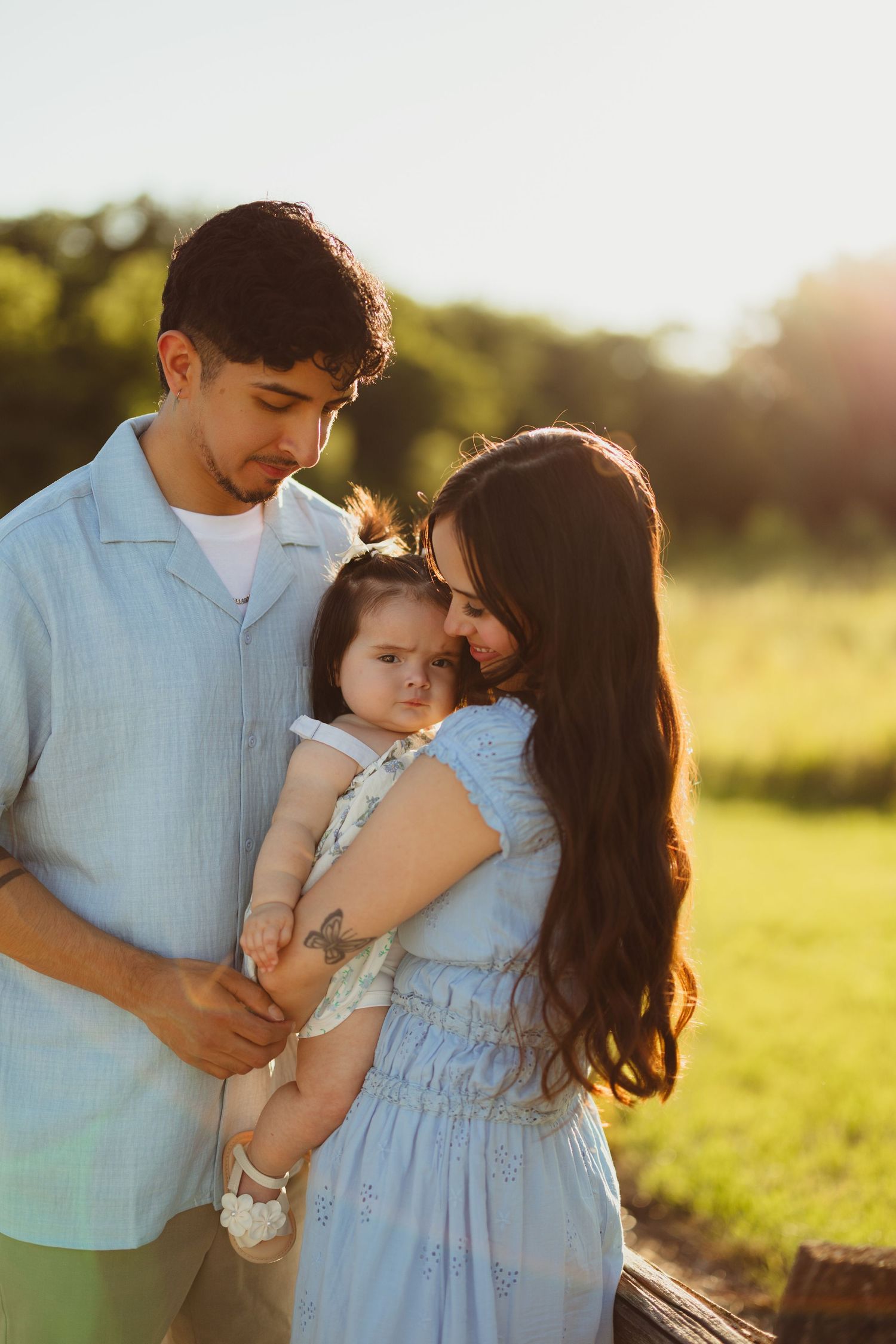 Young family with baby girl poses outdoors in golden hour sunlight, parents in matching blue outfits on green field.
