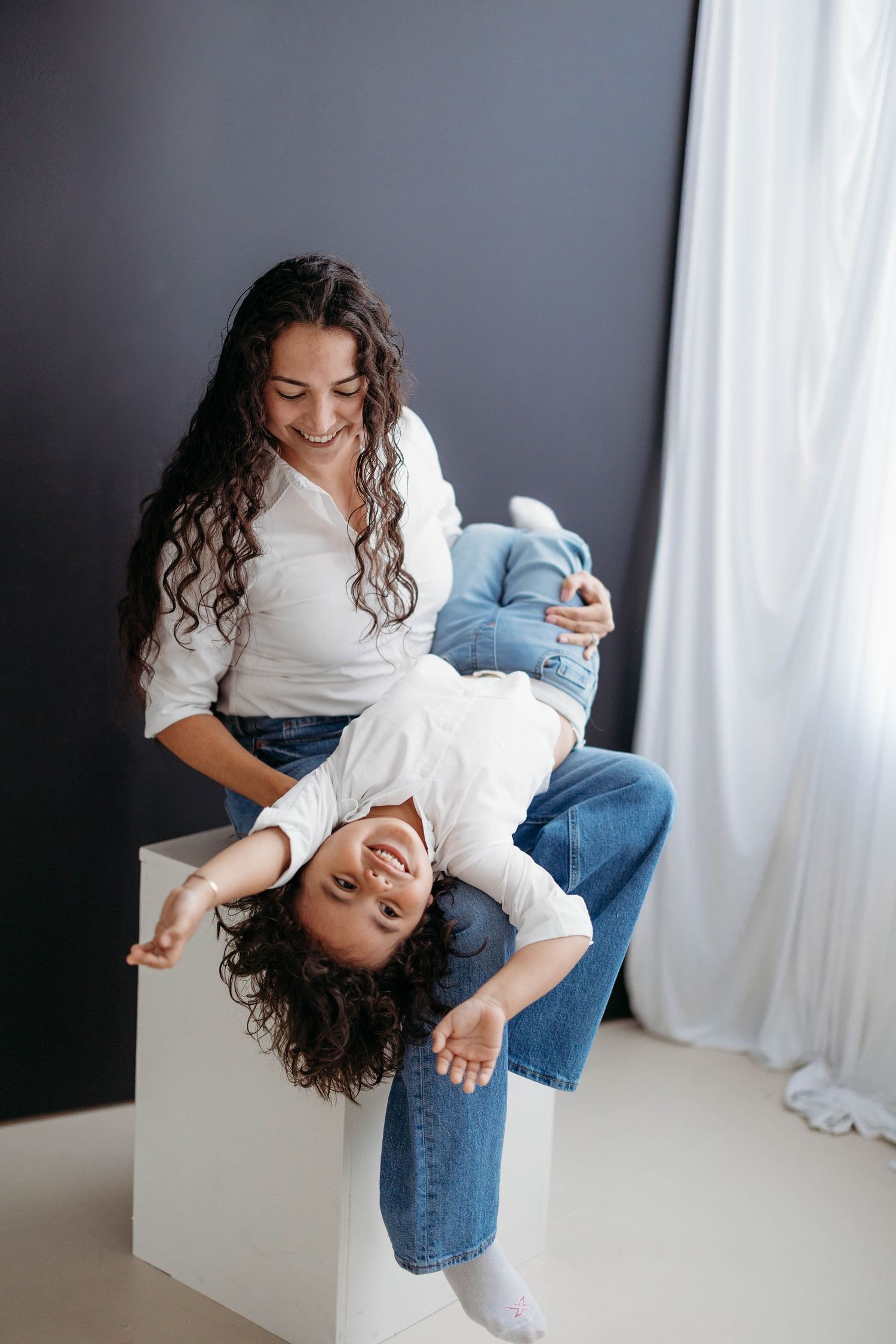 Playful mother holding child upside down while both laugh joyfully in a bright studio. Lafayette indiana tippecanoe county west lafayette