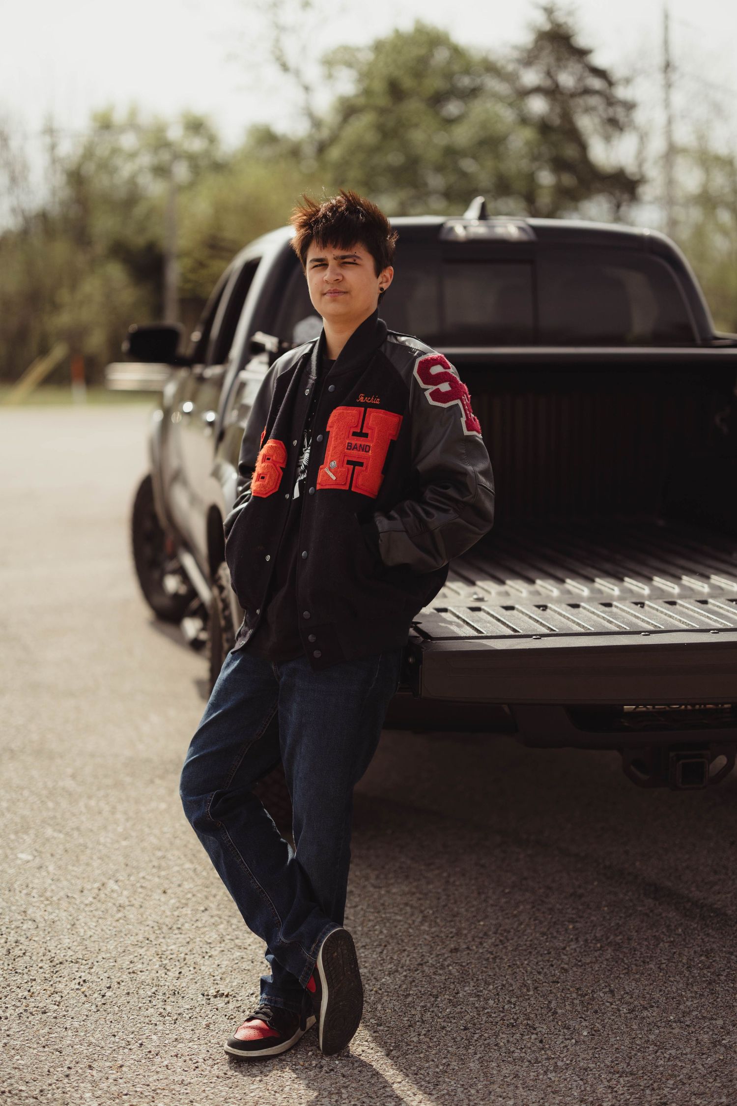 Young man in a varsity jacket leaning against a black pickup truck in a parking lot. High school senior photos in west Lafayette indiana