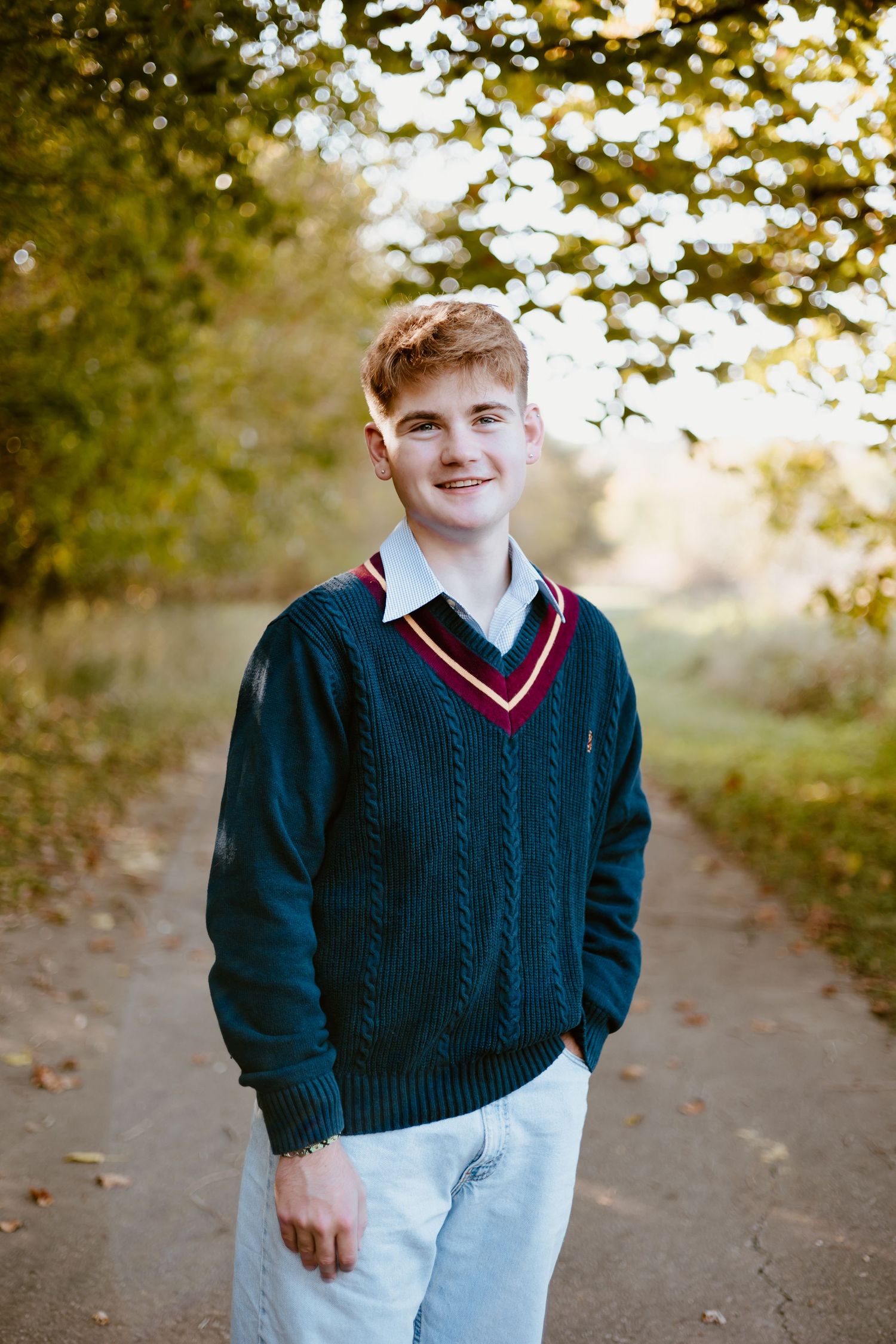 Smiling teenage boy in a dark green V-neck sweater standing on a tree-lined autumn path. High school senior photos in west Lafayette indiana