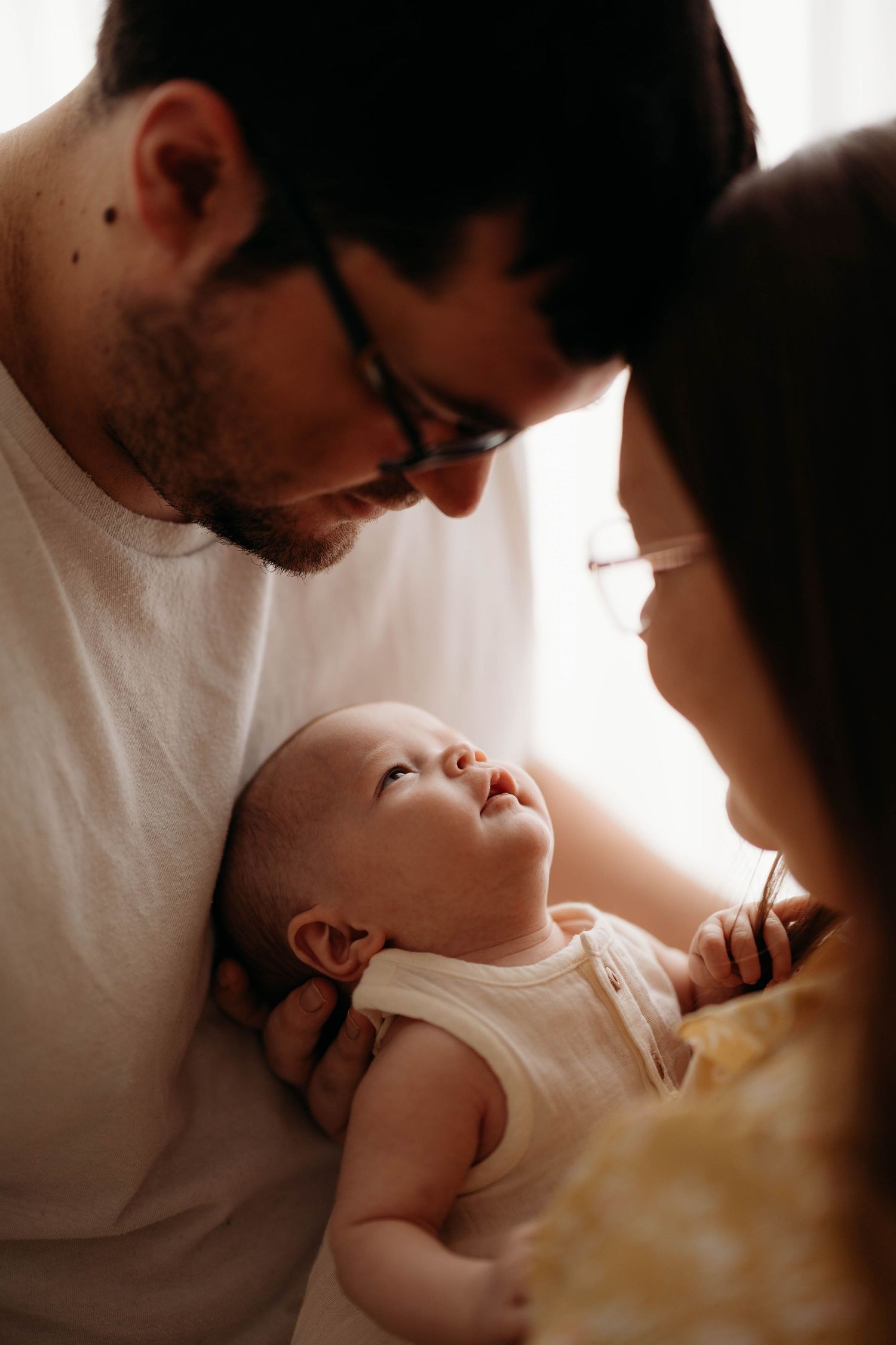 Father with glasses and mother gazing lovingly at alert newborn baby in soft window light indoors. Lafayette indiana tippecanoe county