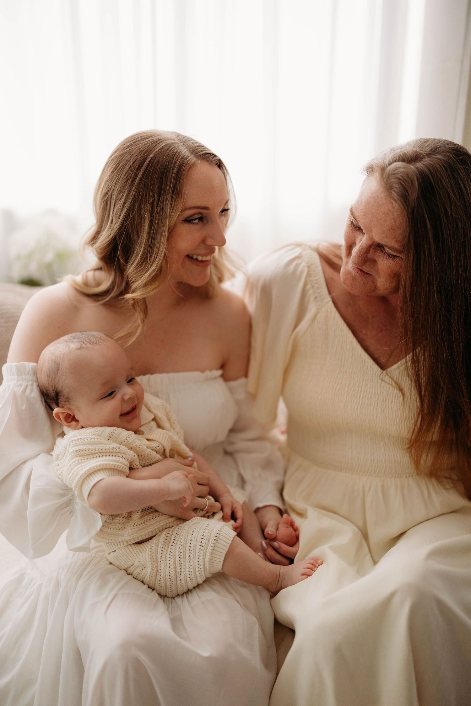 Three generations smile together as young mother holds baby in cream knit outfit on sofa. grandma mom and baby Lafayette indiana