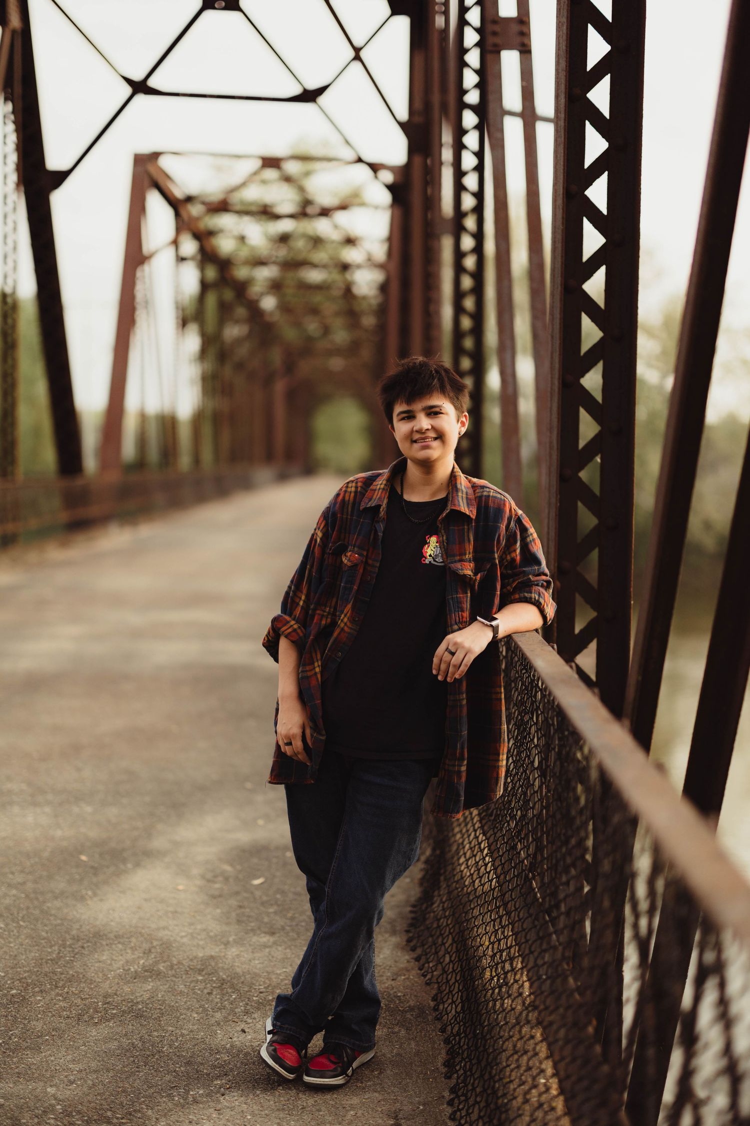 Smiling young man in plaid shirt leaning on a rustic iron bridge railing outdoors. High school senior photos in west Lafayette indiana
