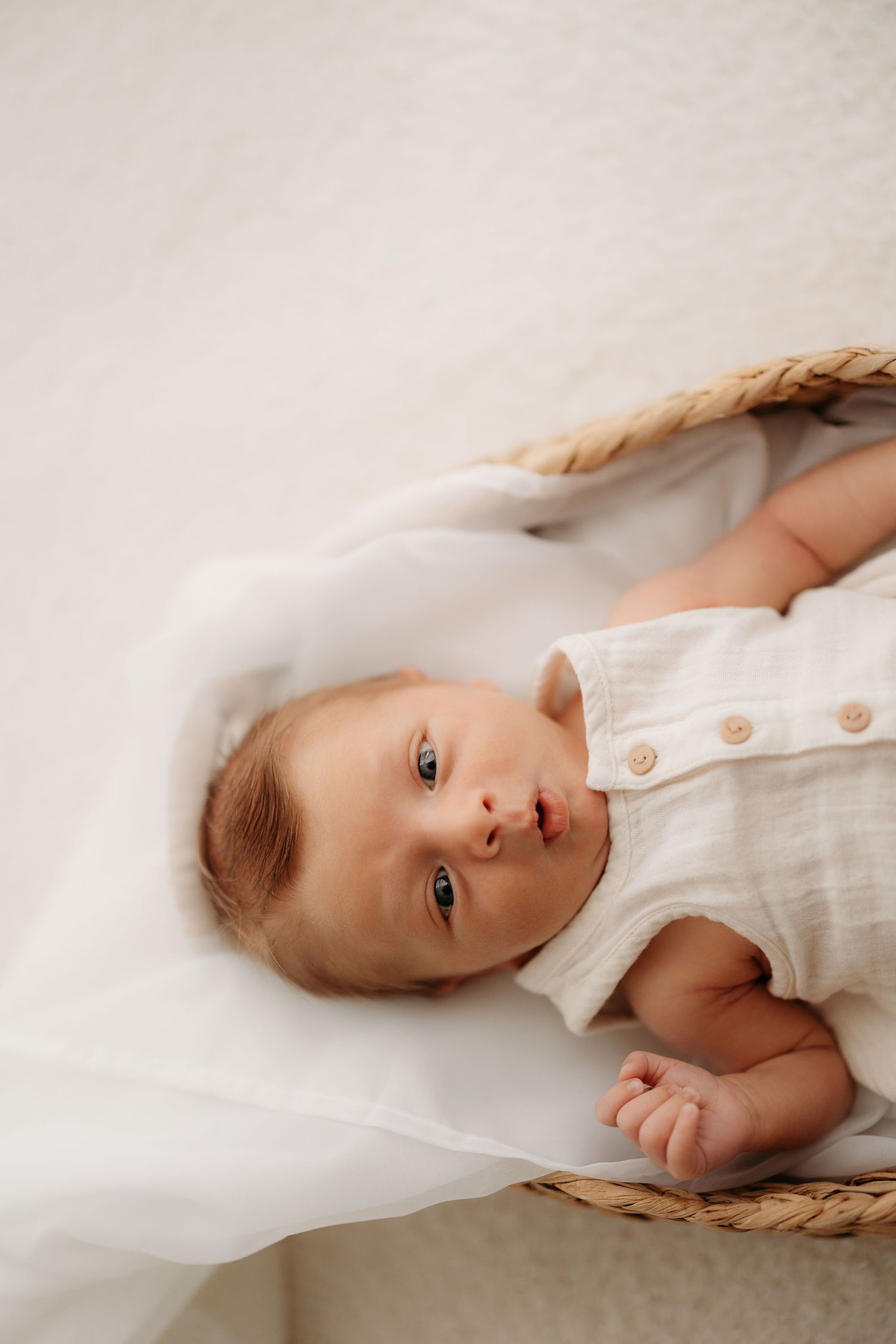 A newborn baby in a white linen romper lying in a woven basket with soft white fabric.