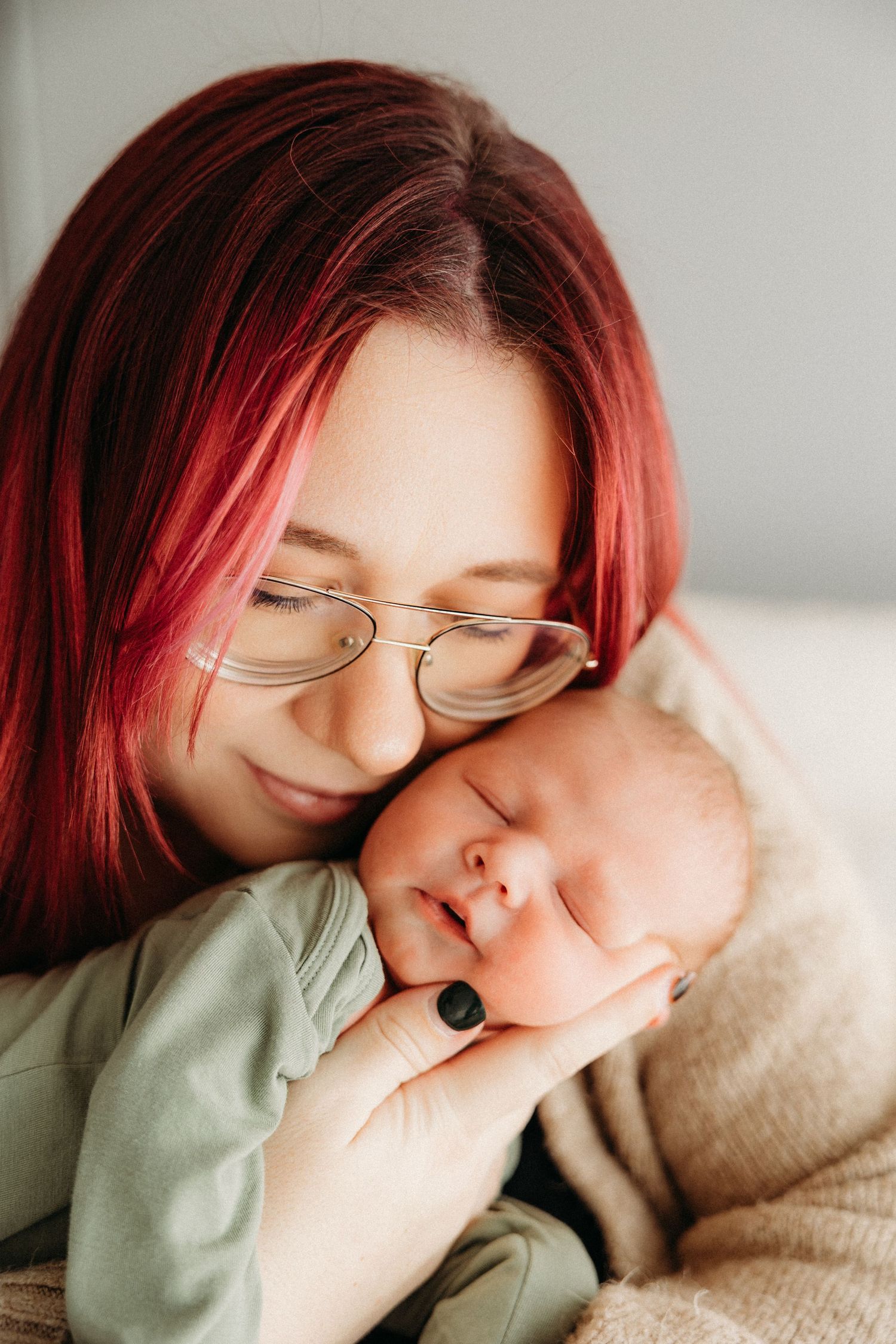 Red-haired woman with glasses tenderly holds sleeping newborn baby close to her face.