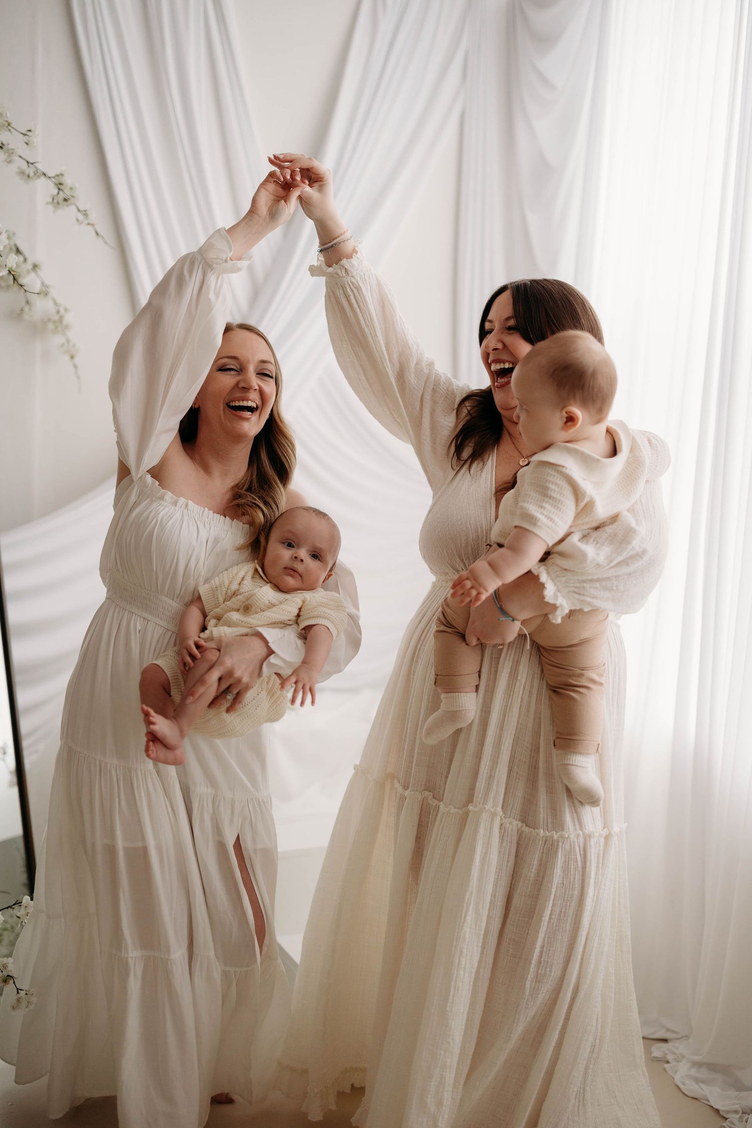 Two laughing mothers in white dresses hold their babies while dancing together in a bright studio. best friend motherhood photos