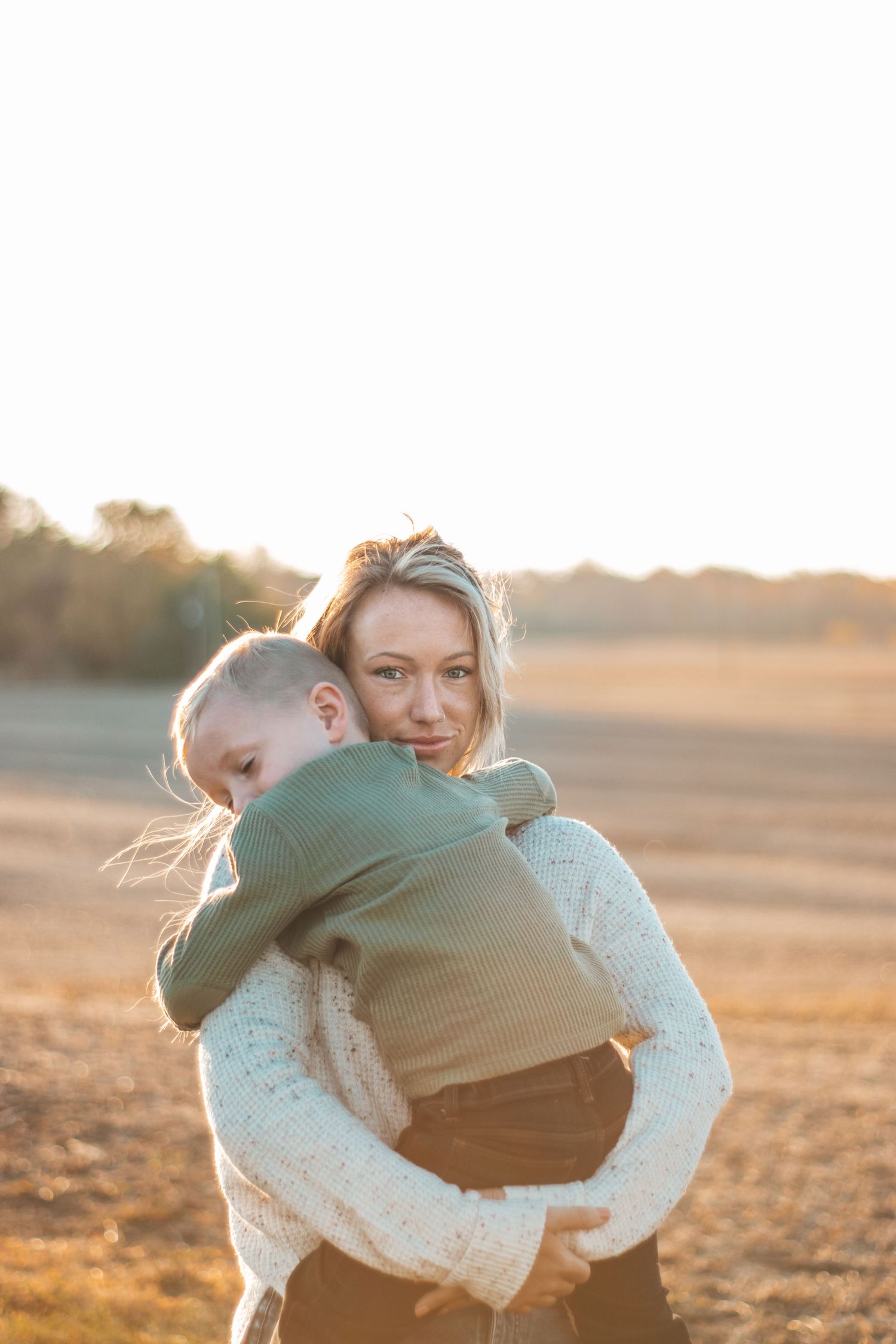 Blonde mother holding young child in warm golden sunset light in open field.