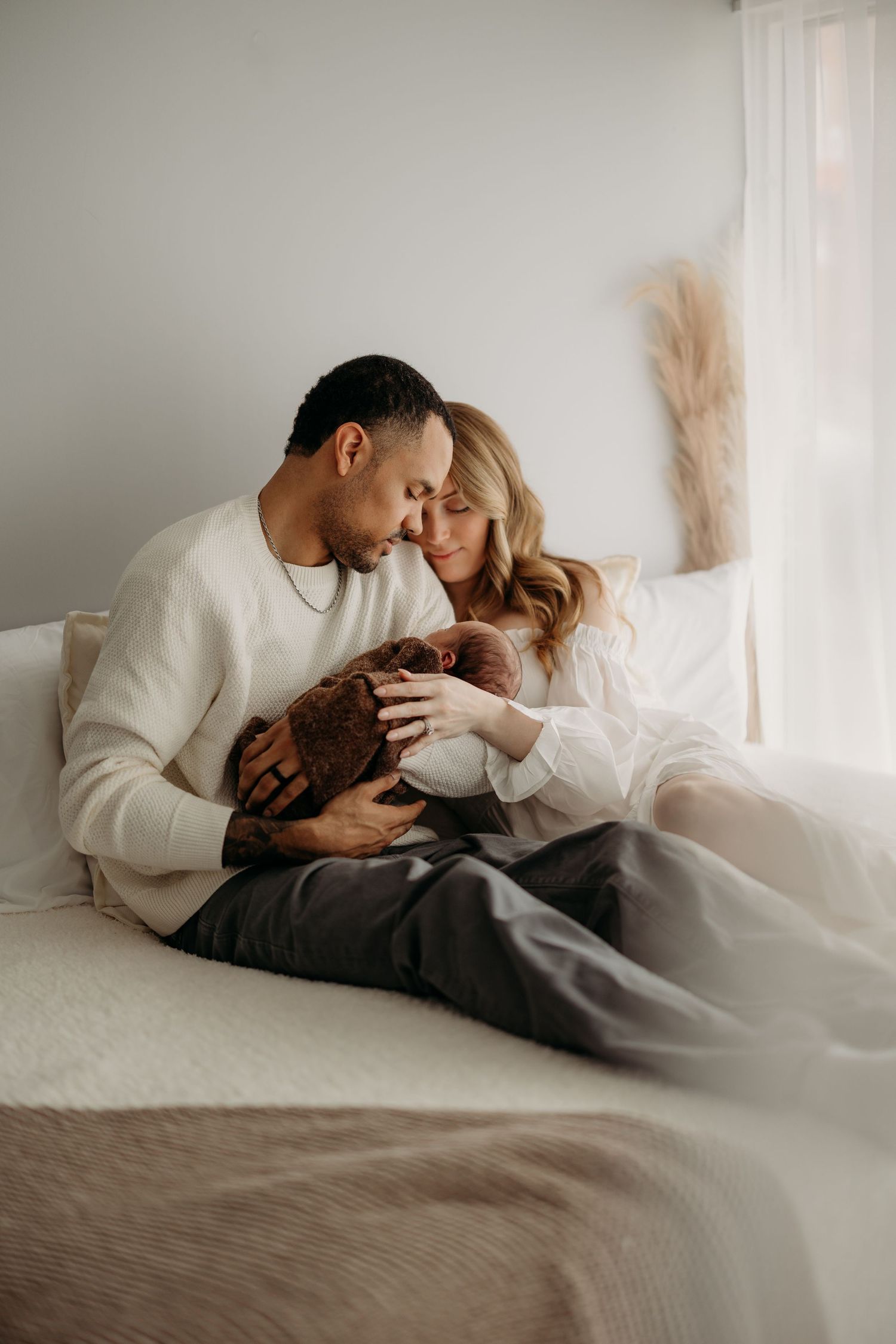 A couple sits on a bed, lovingly cradling their newborn baby in a soft, neutral-toned room.