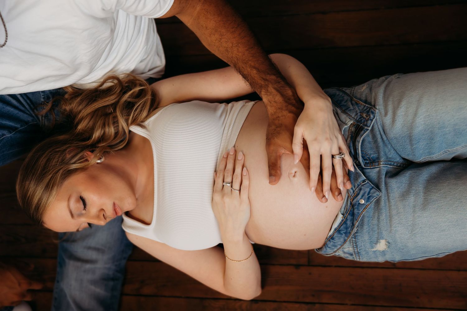 A pregnant woman in a white top and jeans lies on a wooden floor with a partner. Lafayette indiana tippecanoe county west lafayette
