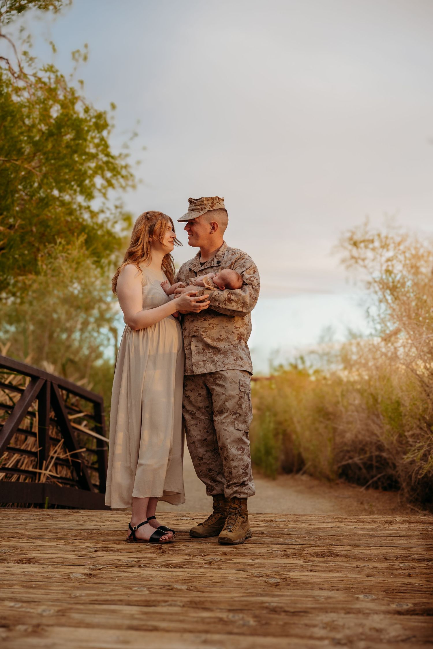 Military couple in desert at sunset, soldier in camouflage holding newborn while woman in white dress stands close. Lafayette indiana