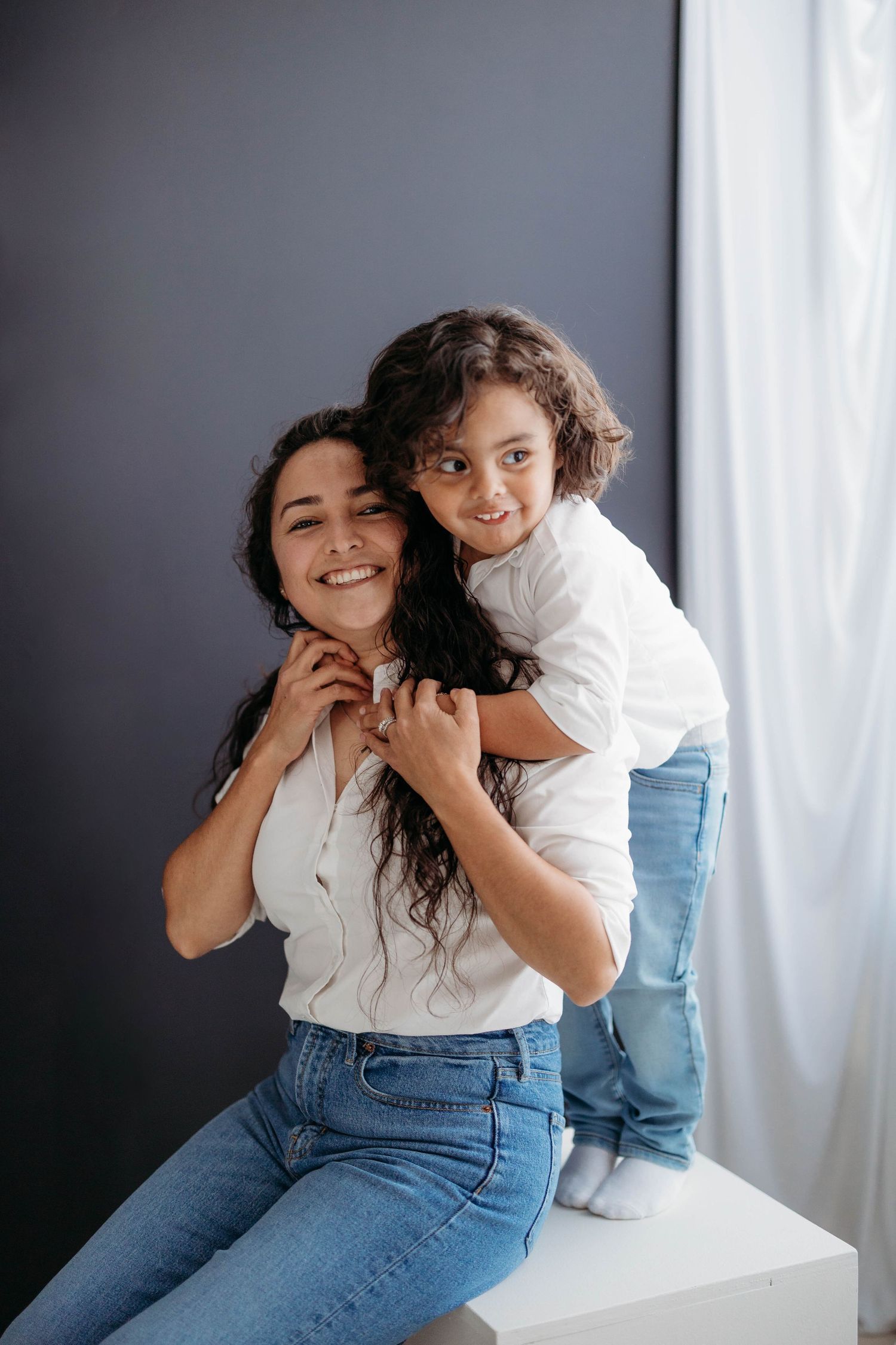 Smiling mother in white shirt and jeans hugged from behind by young child in studio. Lafayette indiana tippecanoe county west lafayette