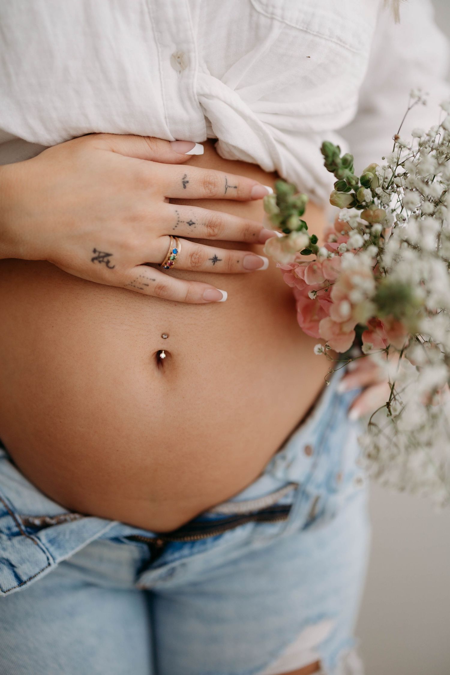 Close-up of pregnant belly with tattooed hands holding bump, wearing jeans and white shirt with flowers. maternity pregnancy announcement