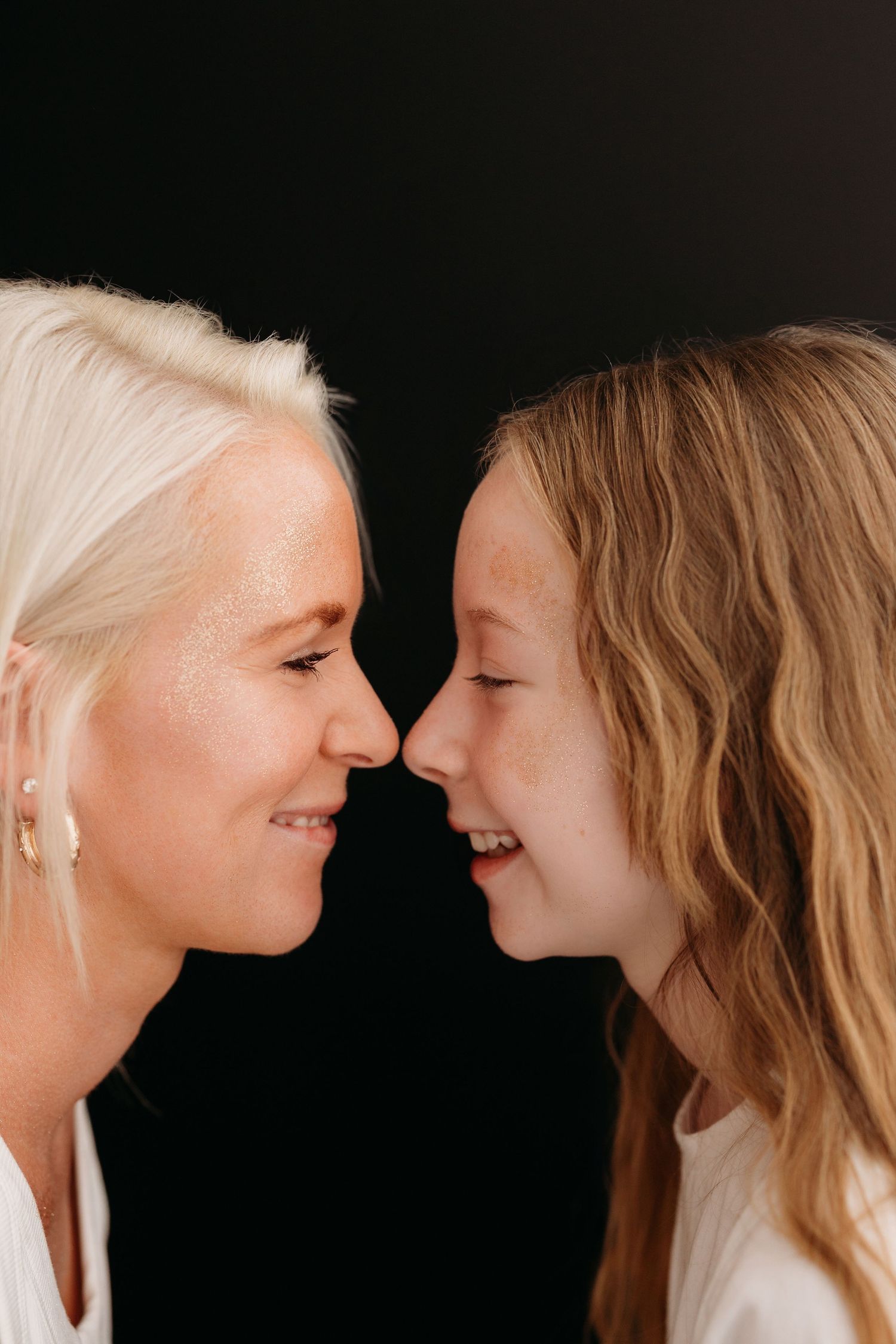 A blonde woman and young girl smile facing each other, touching noses against a black background.
