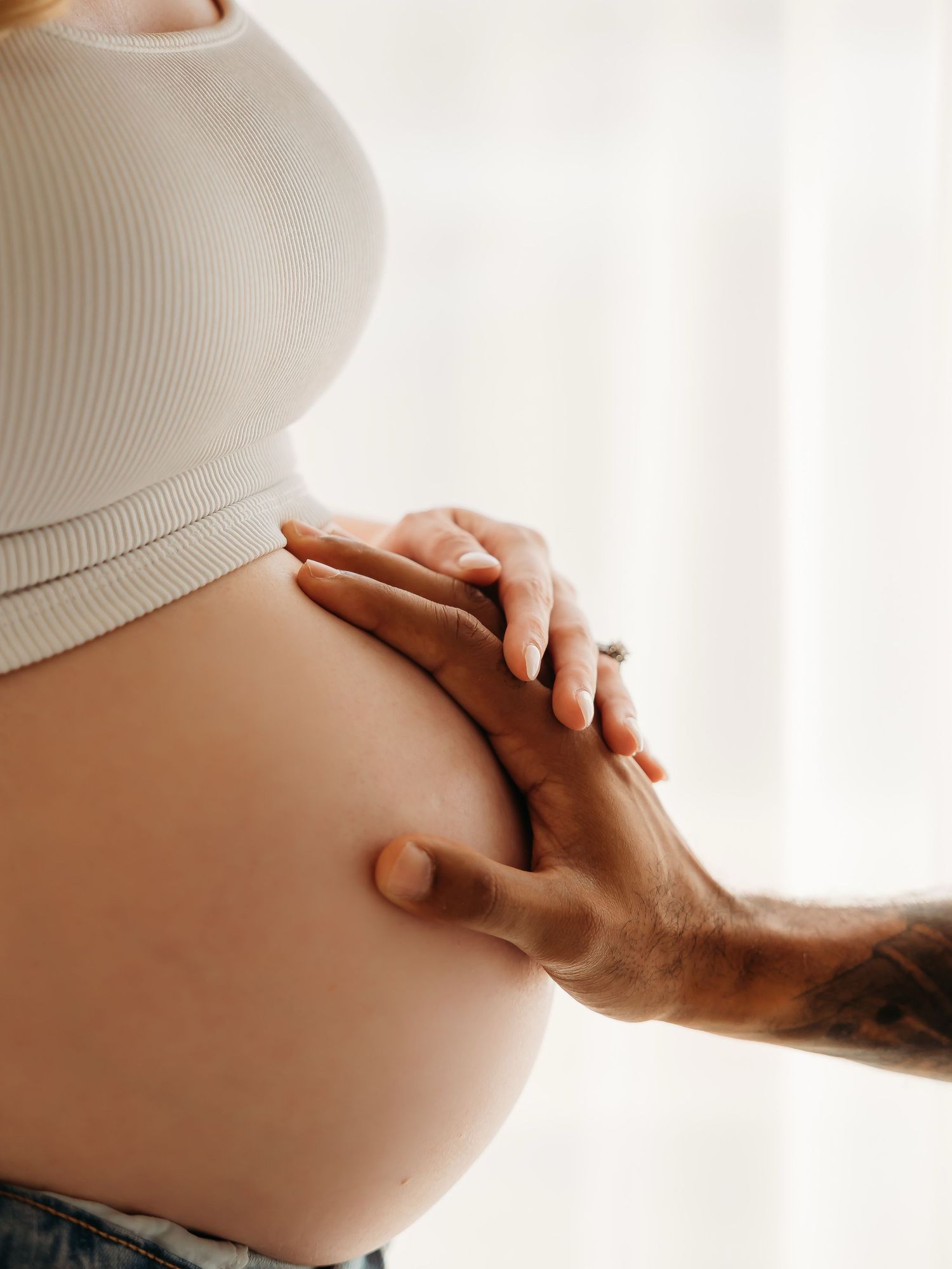 Pregnant woman in white crop top with partner's tattooed hand gently touching her baby bump. Lafayette indiana tippecanoe county