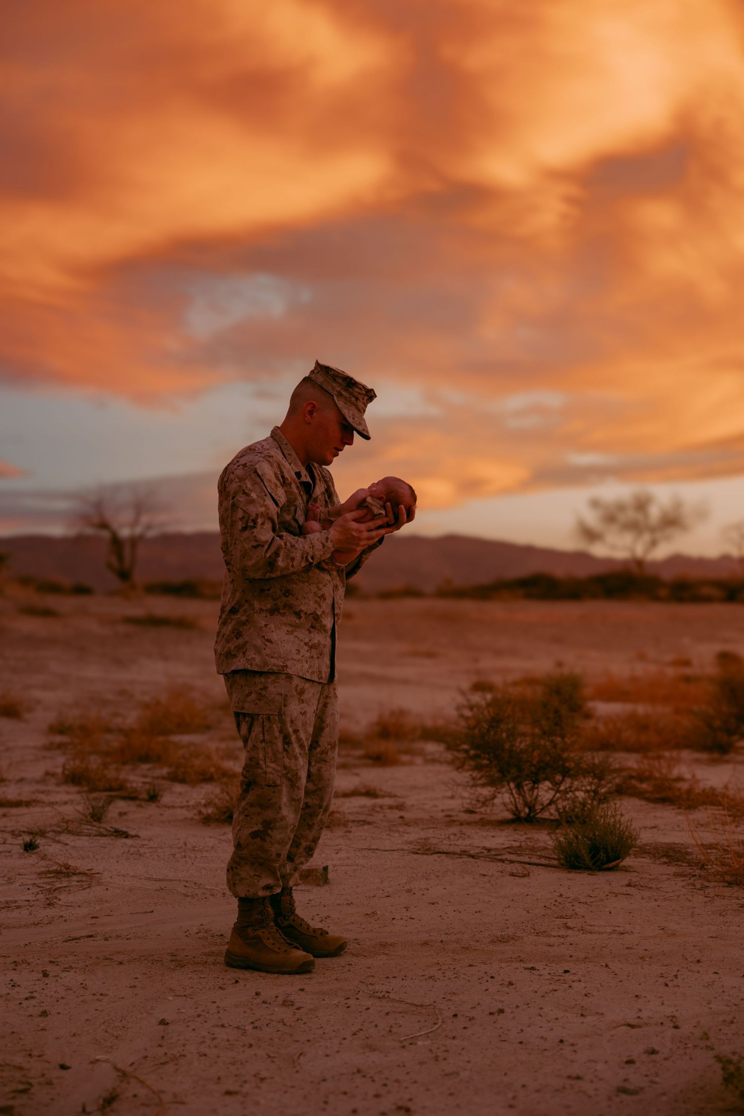 US Marine soldier in camouflage uniform holding newborn baby at sunset in desert landscape with dramatic orange sky. Lafayette indiana
