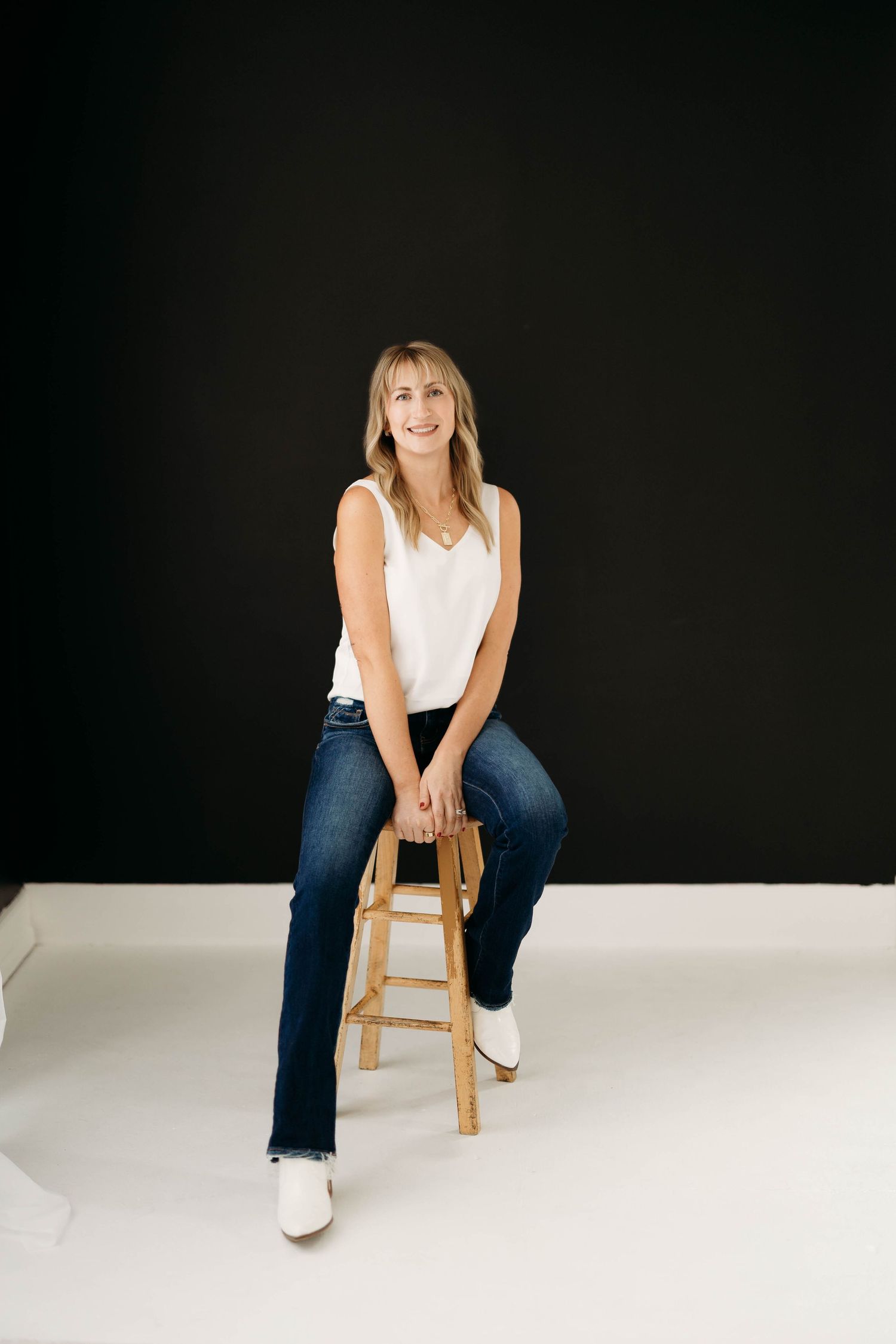 Smiling blonde woman in white tank top and dark jeans sitting on wooden stool against black background. Branding photos in west Lafayette In
