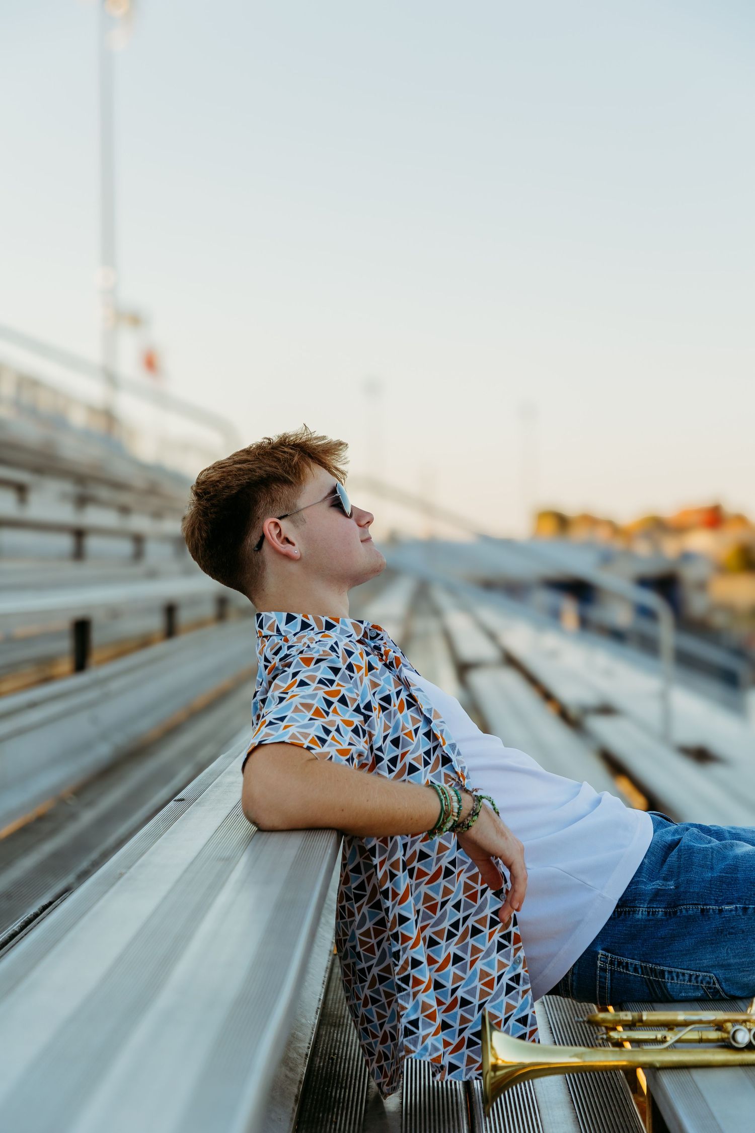 Teen boy in patterned shirt on bleachers at sunset with a band instrument nearby. High school senior photos in west Lafayette indiana