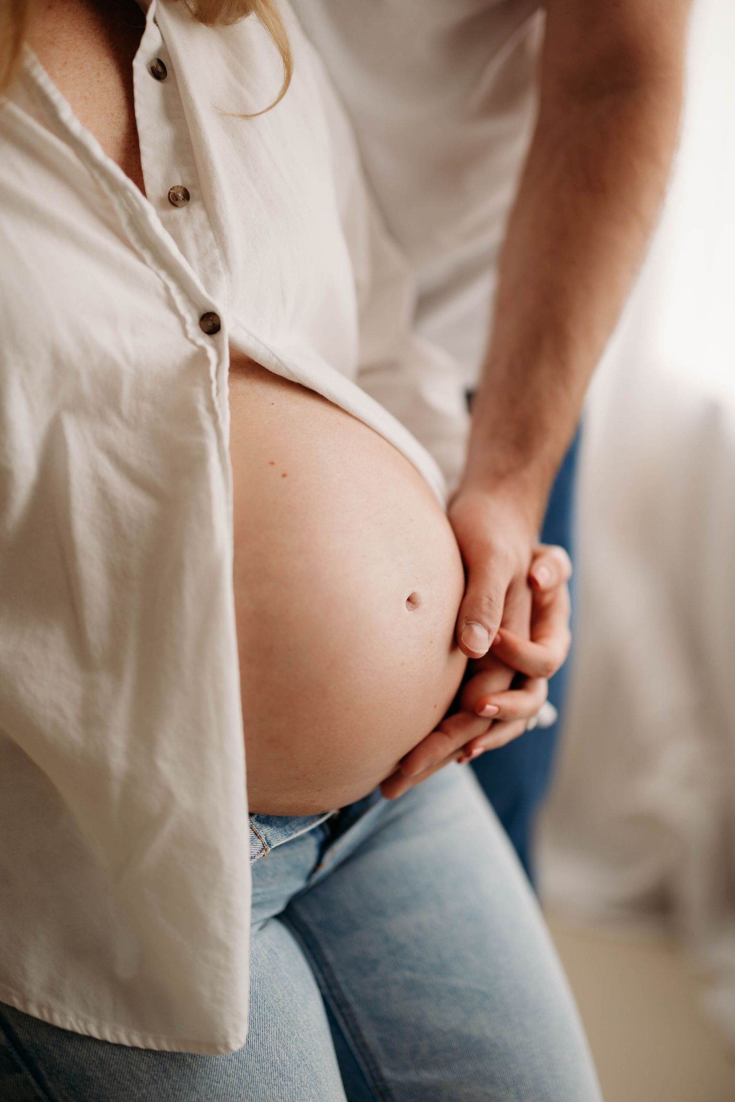 Pregnant woman in white shirt and jeans cradling baby bump with partner's hand gently resting on belly.
