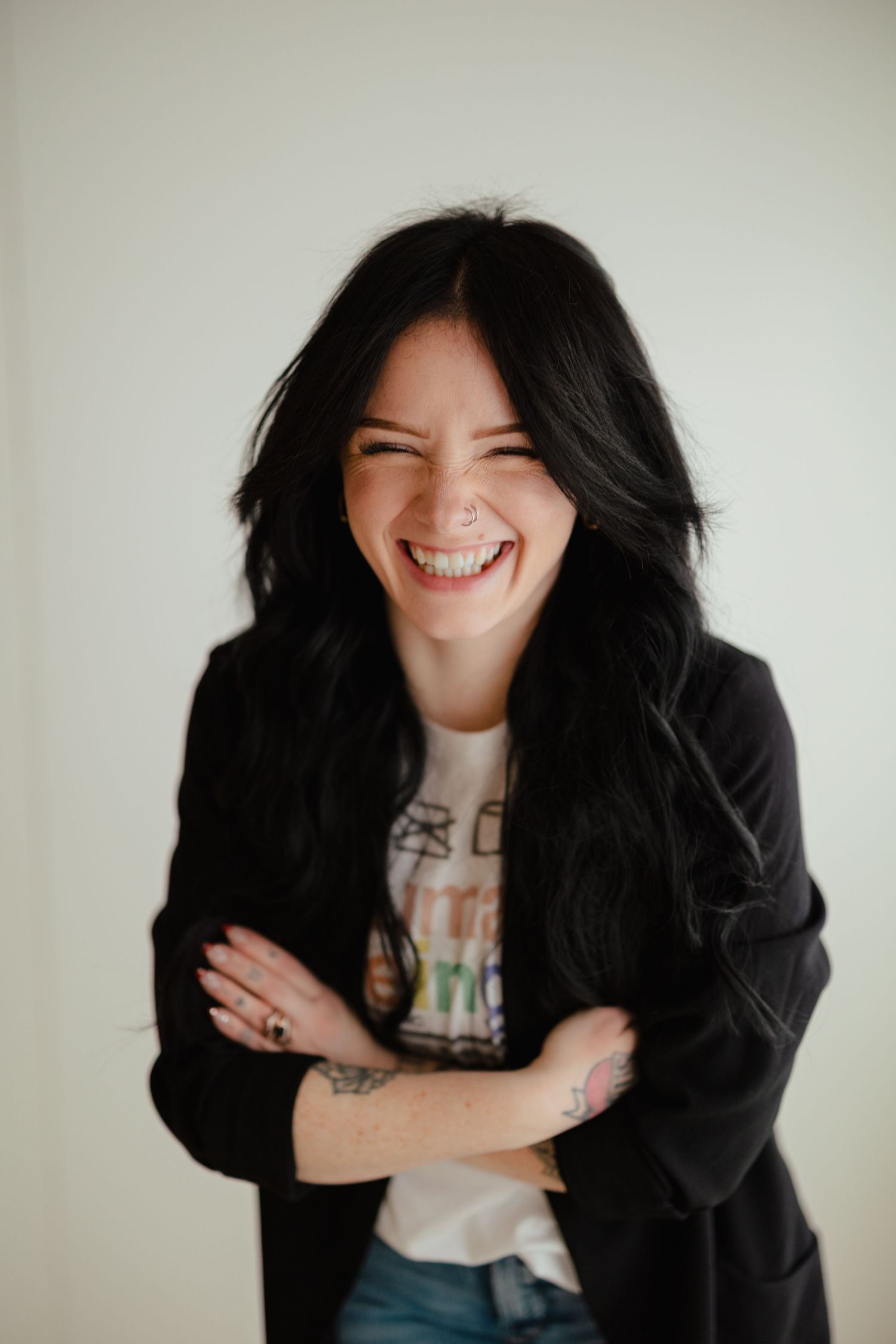 Laughing dark-haired woman with tattoos in black blazer and graphic tee against white wall. Branding photos in west Lafayette indiana