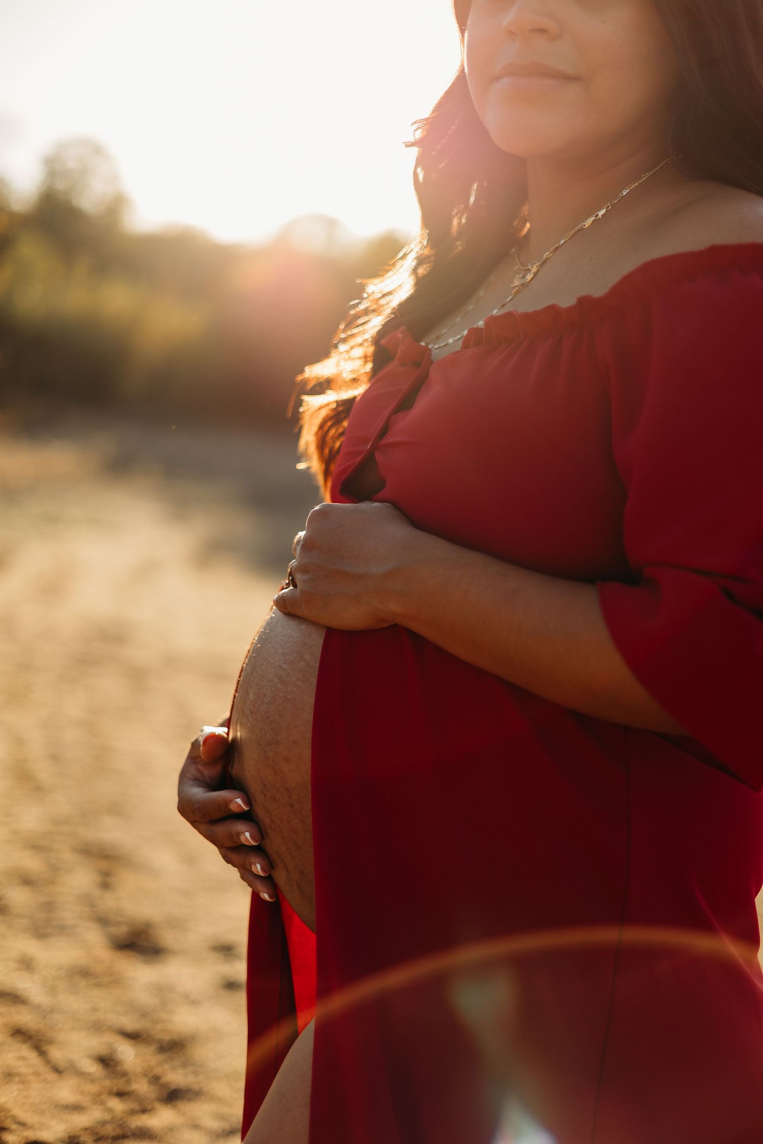 Close-up of pregnant woman in red off-shoulder dress holding bump in golden sunset light outdoors. Lafayette indiana tippecanoe maternity