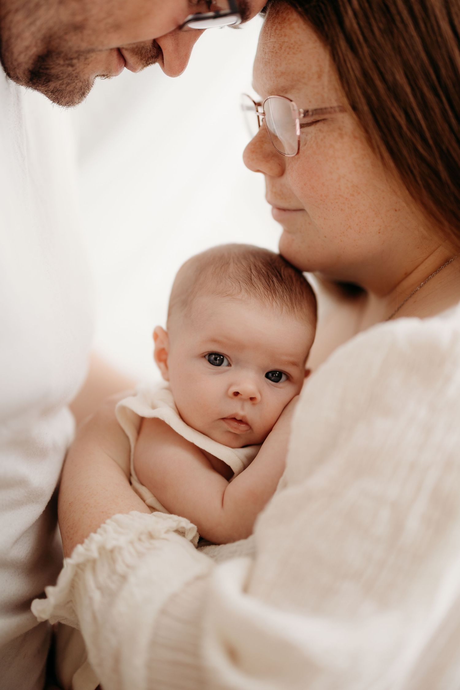 Loving parents with beards and glasses gently cradle alert newborn baby dressed in cream outfit. Lafayette indiana tippecanoe county