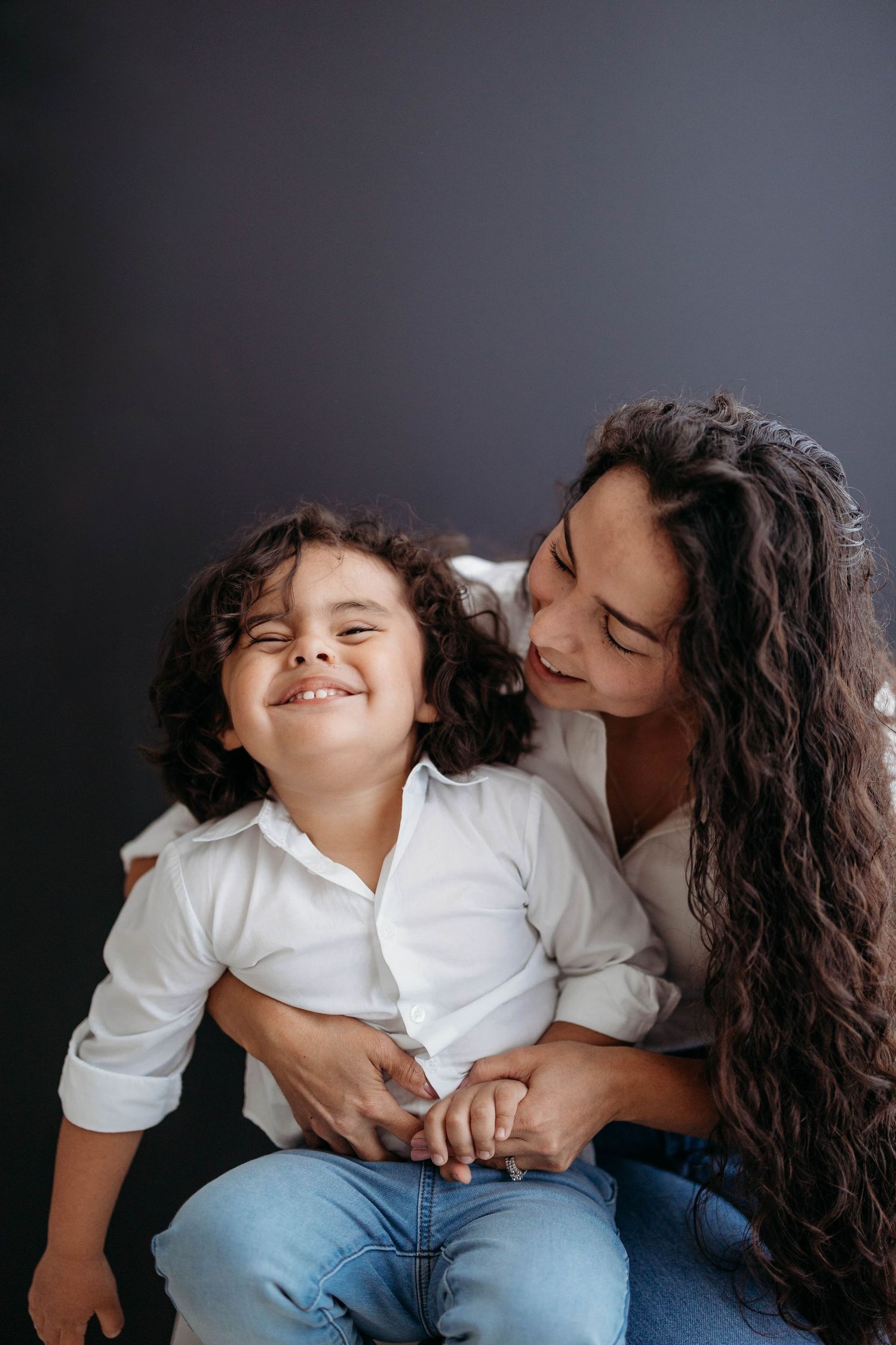 Joyful mother in white shirt hugging her laughing curly-haired child against dark background. Lafayette indiana tippecanoe county west lafay