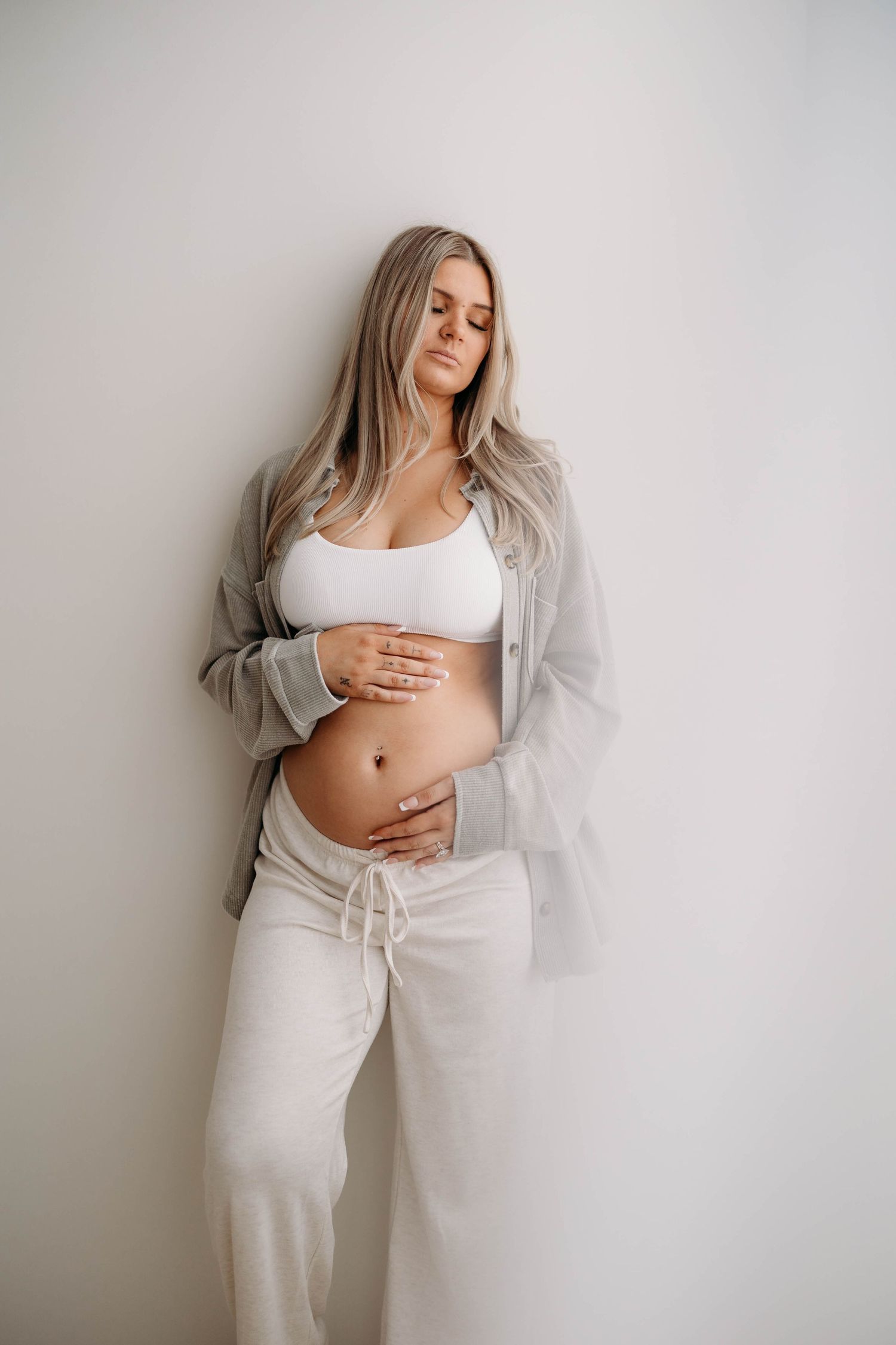 Blonde pregnant woman in white bra and grey shirt cradling bare bump against wall. Lafayette indiana tippecanoe county west lafayette