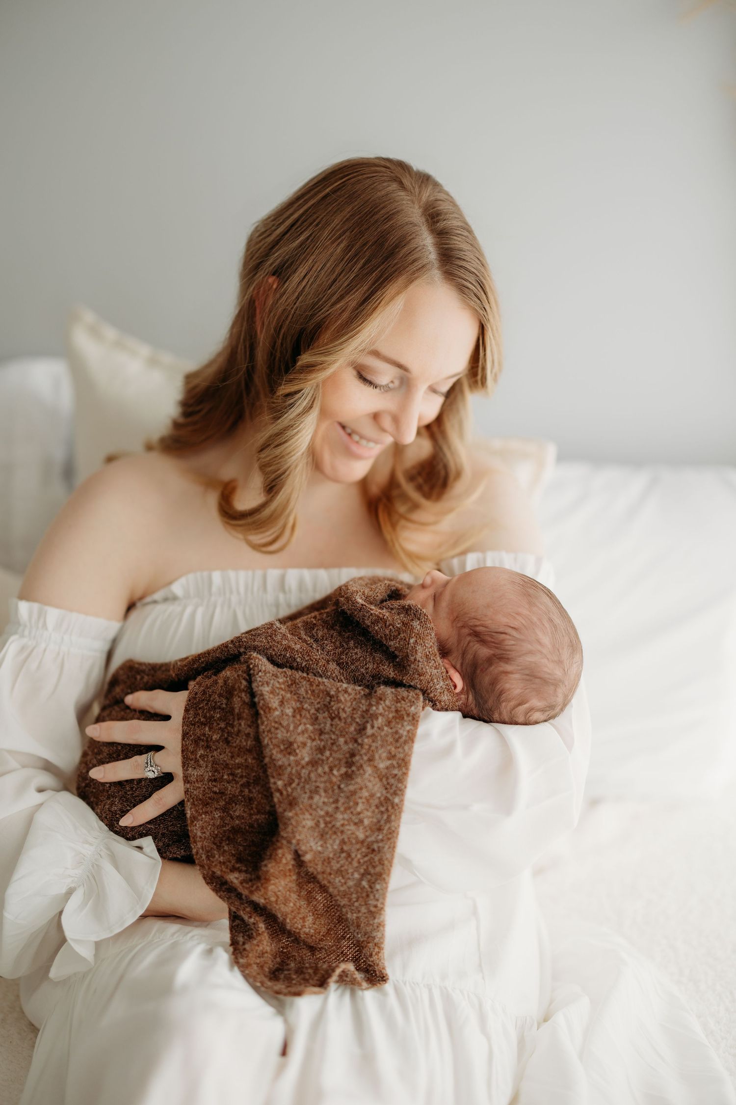 Smiling mother in white dress tenderly holds sleeping newborn wrapped in brown knit blanket on white bed.
