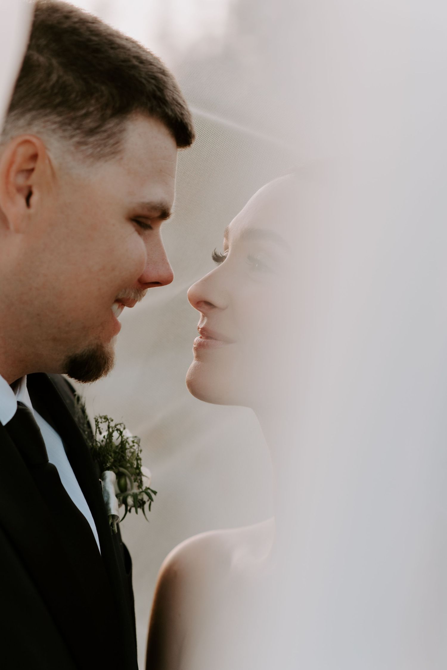 A bride and groom share an intimate moment face to face, softly lit through a sheer white wedding veil.