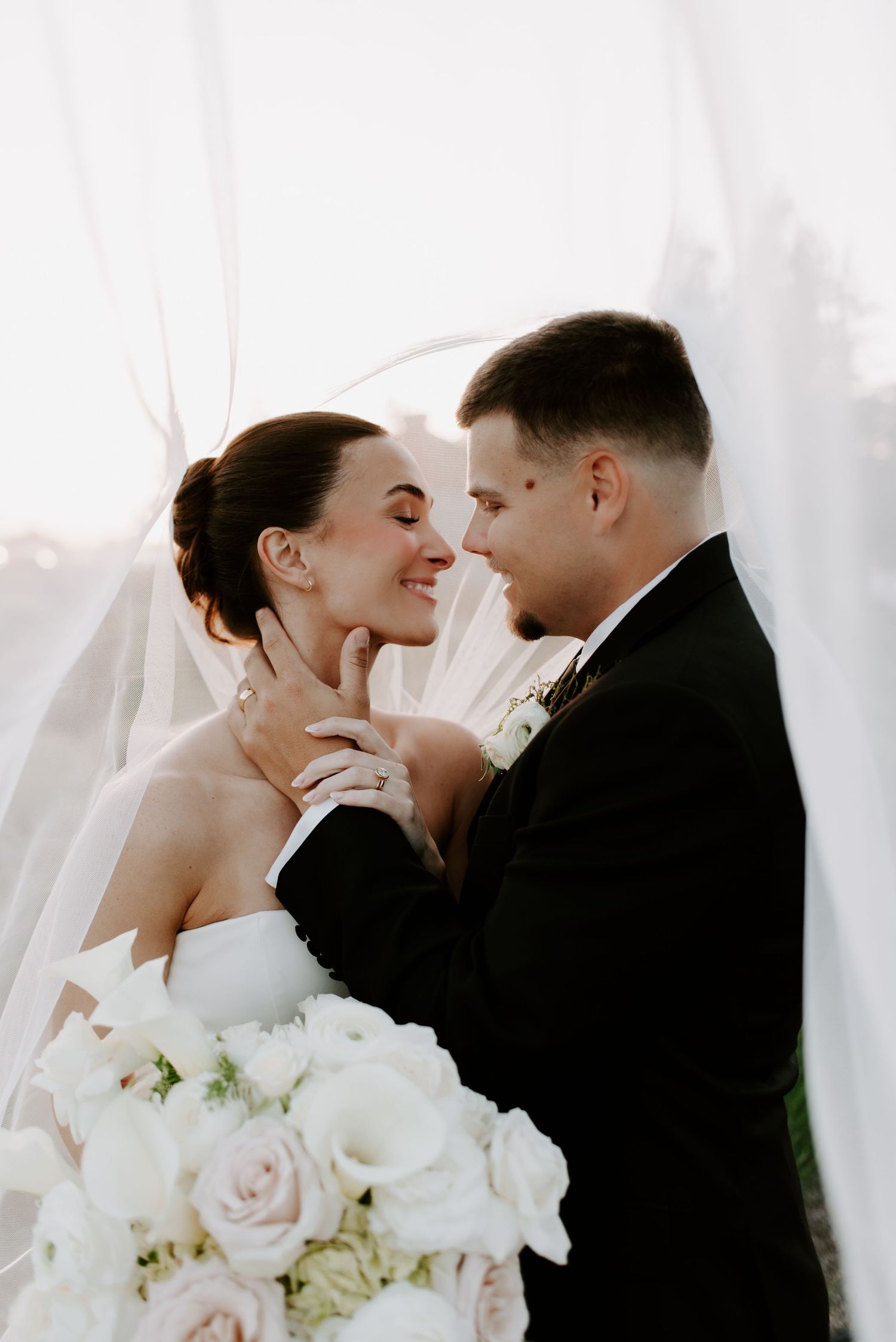 Bride and groom share a romantic moment under a flowing veil, bride holding a white rose bouquet.