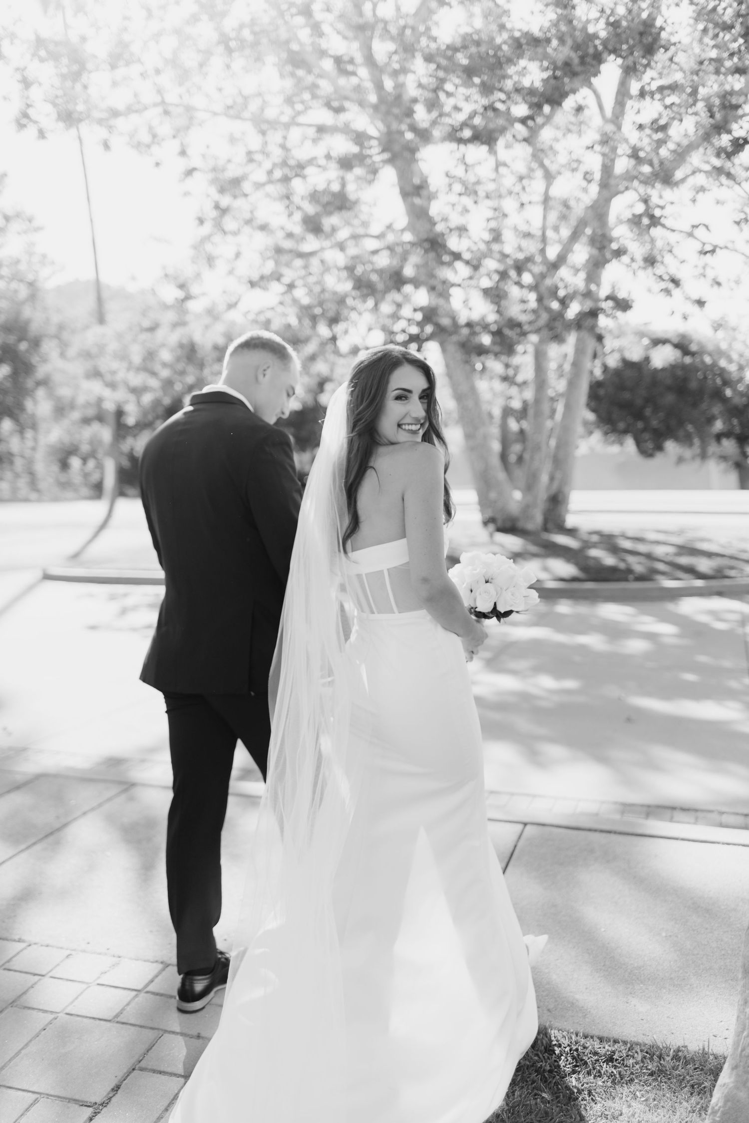 Black and white photo of a smiling bride in a white gown and veil holding bouquet, walking with groom in black suit outdoors.