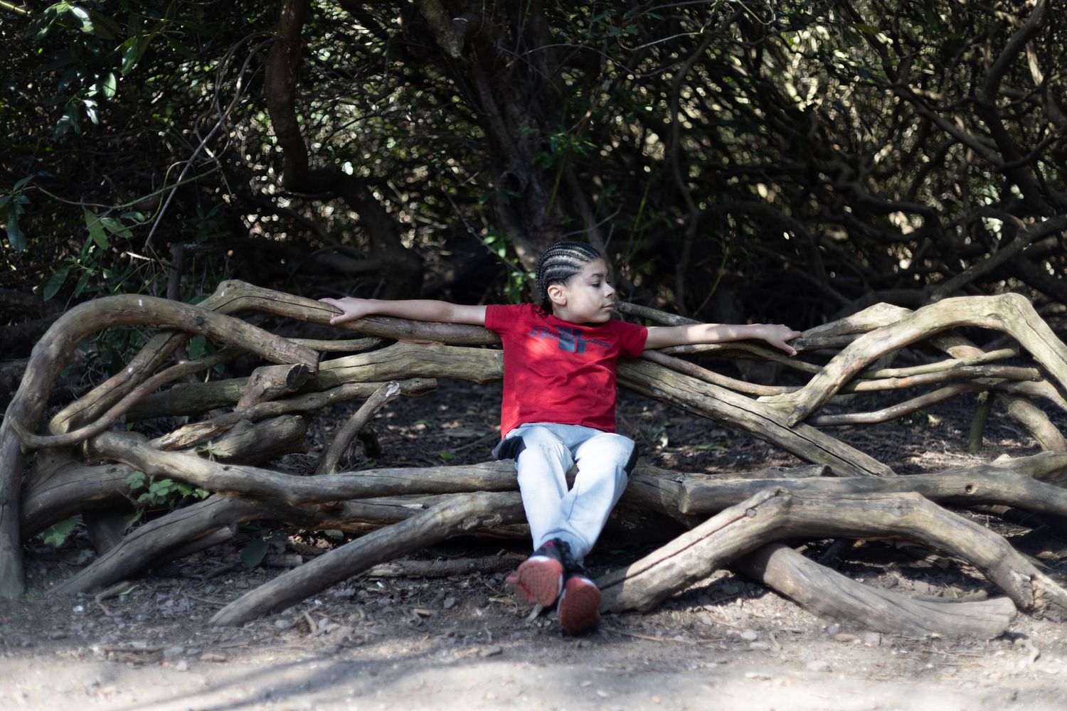 Young child in red shirt sitting relaxed on a pile of logs in a shaded woodland area.