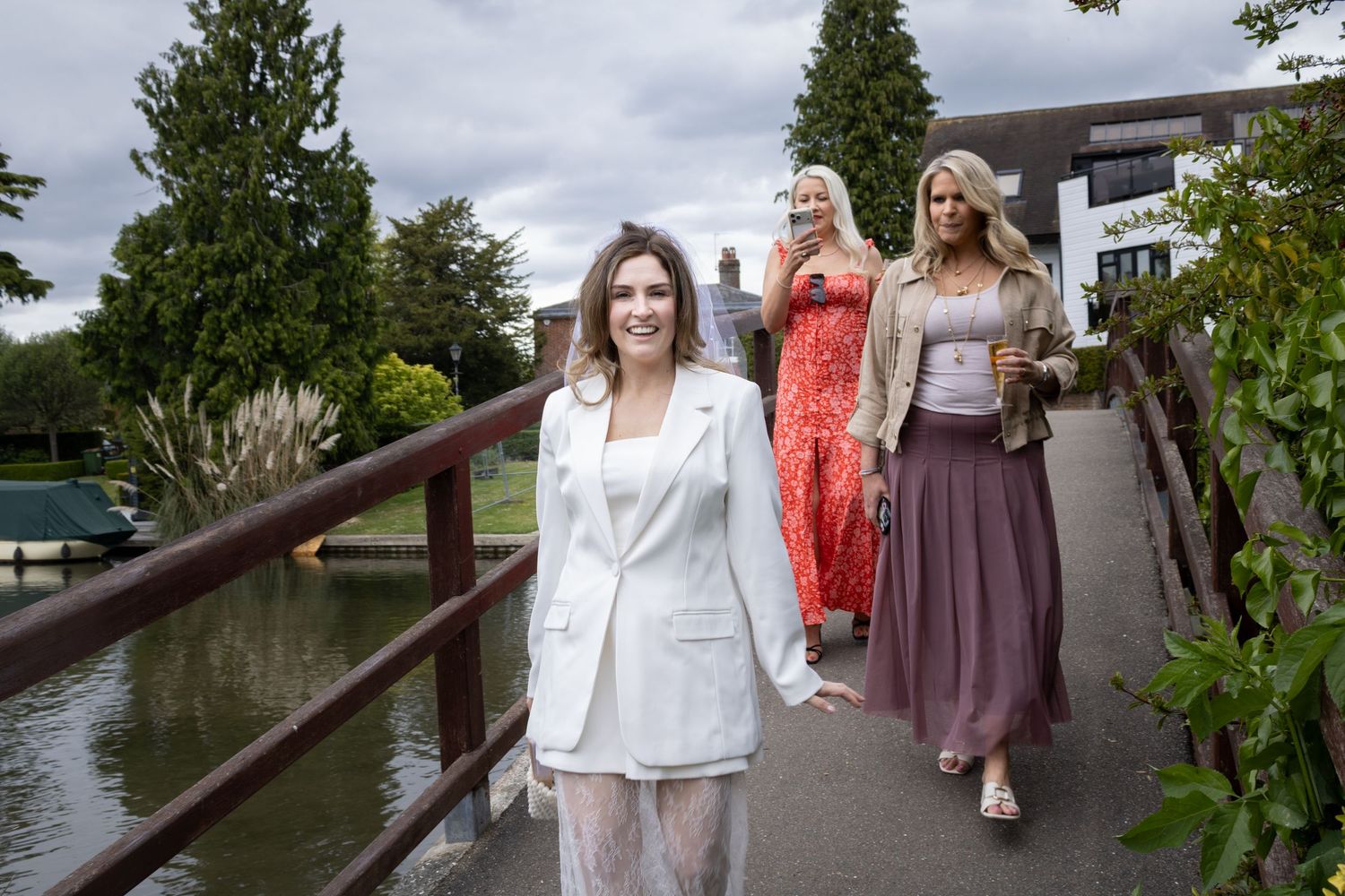 Three women in elegant dresses walking across a bridge near a scenic waterway venue.