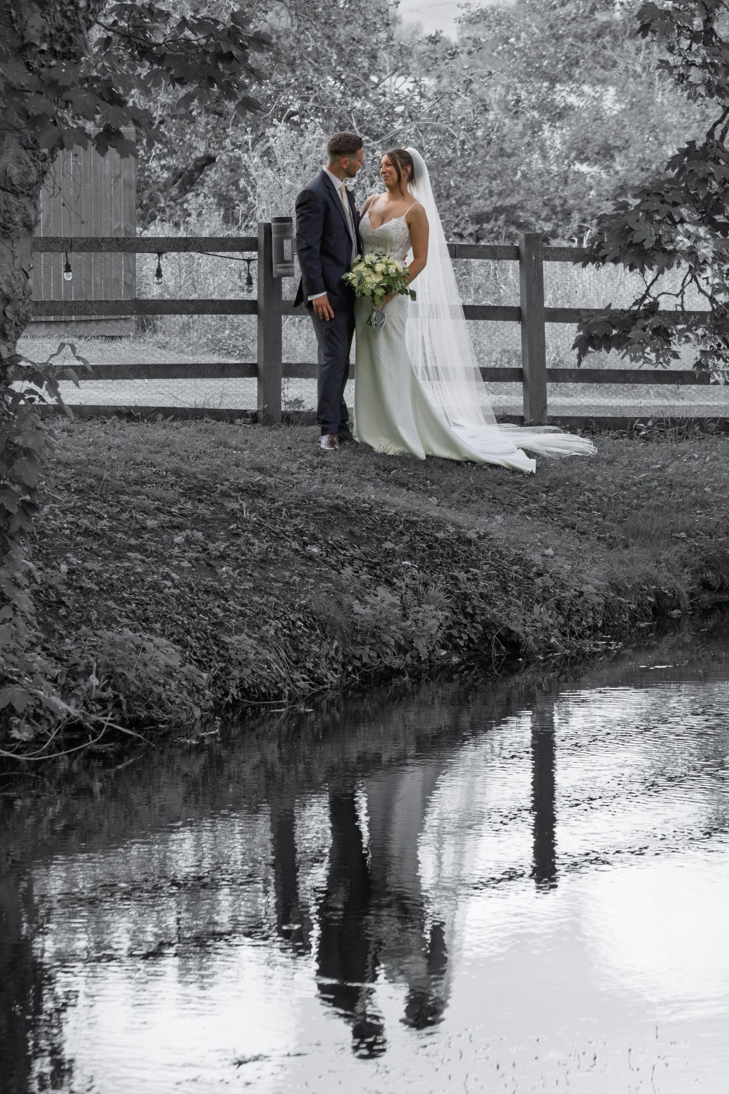 Bride and groom stand by a reflective stream in a black and white wedding photo with a rustic fence backdrop.