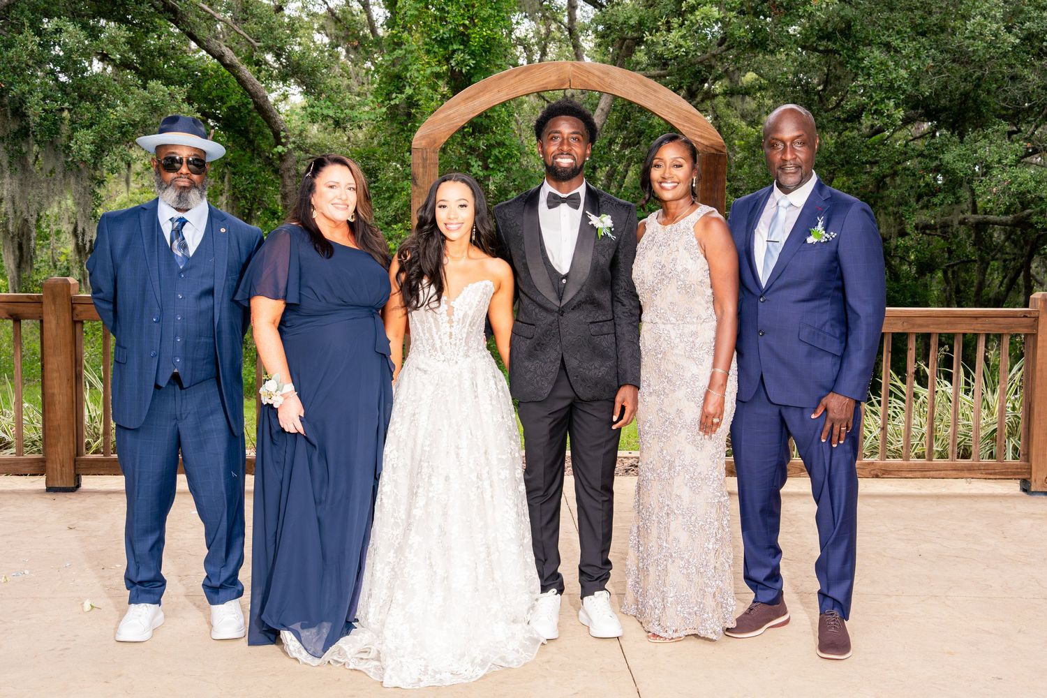 Wedding party of six poses under wooden arch with bride in white gown and groom in tuxedo.